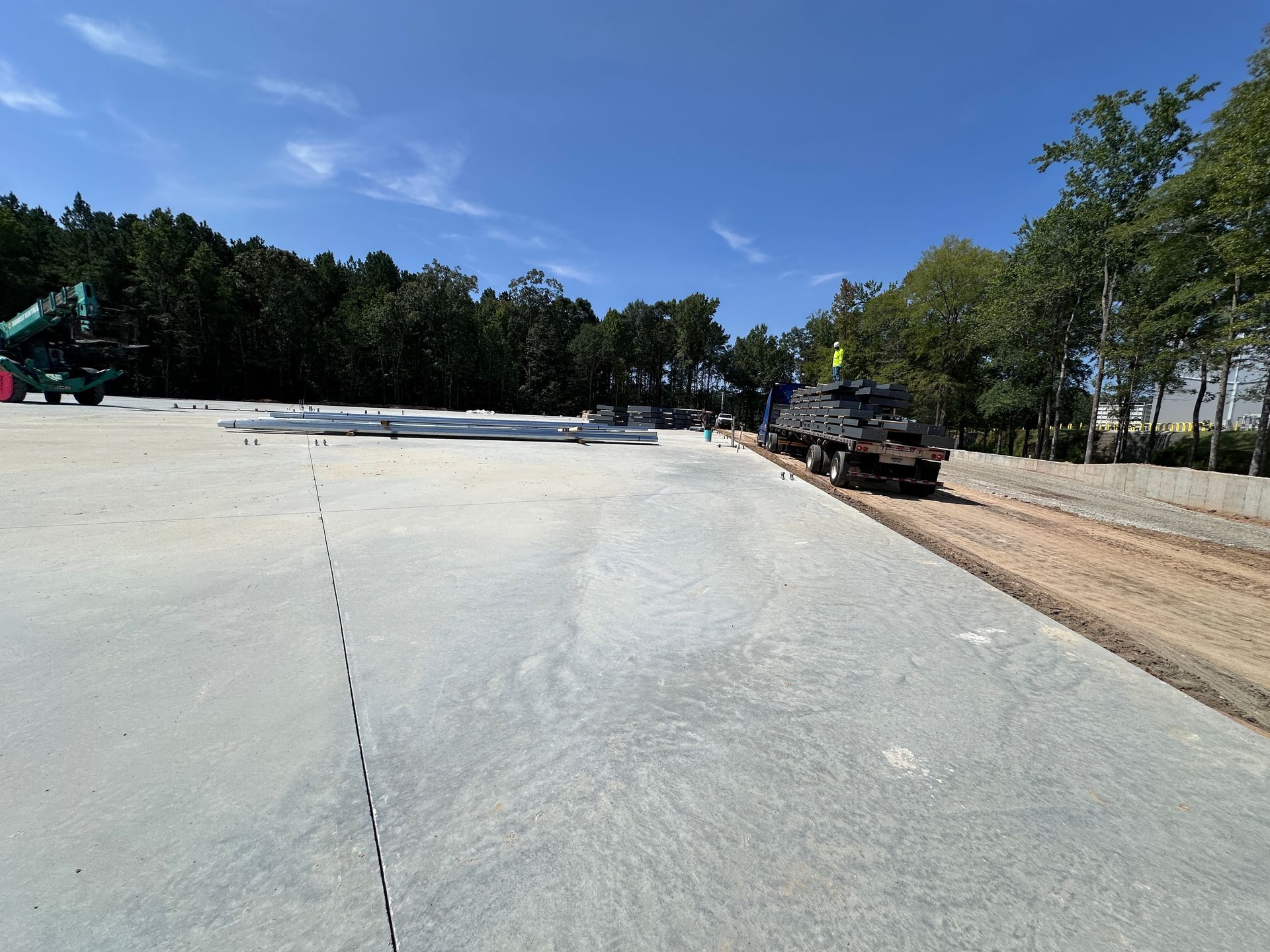 Wide concrete construction site with a blue sky, trees, and excavators in the distance