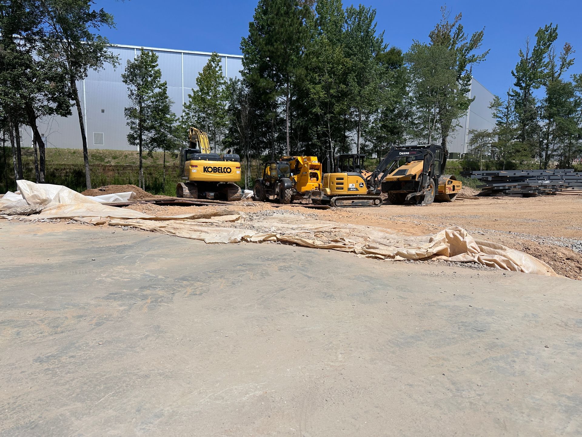 Yellow construction vehicles on a dirt site near trees and a building under a clear blue sky