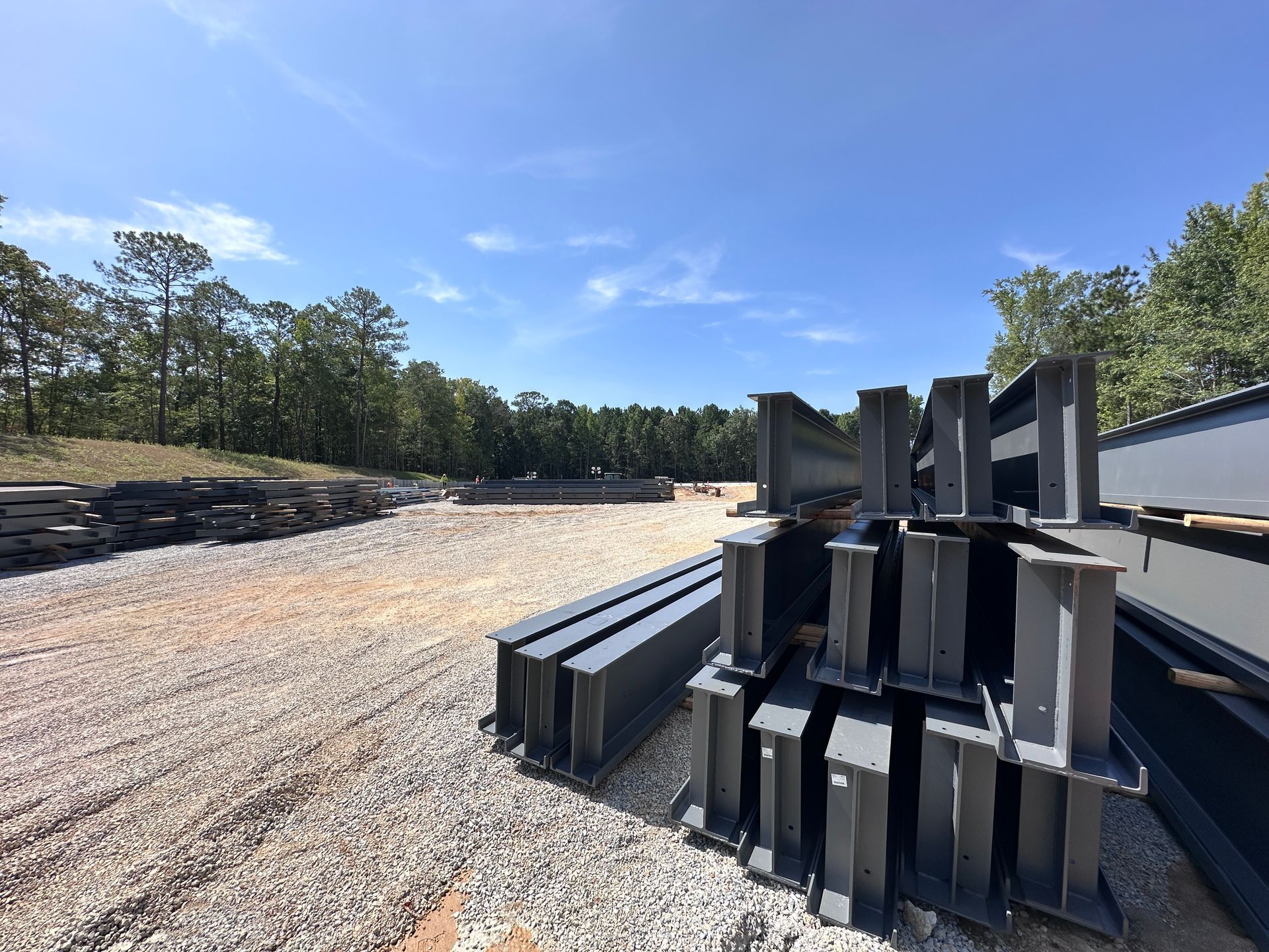 Construction site with dirt piles, heavy equipment, and a tree line under a partly cloudy blue sky