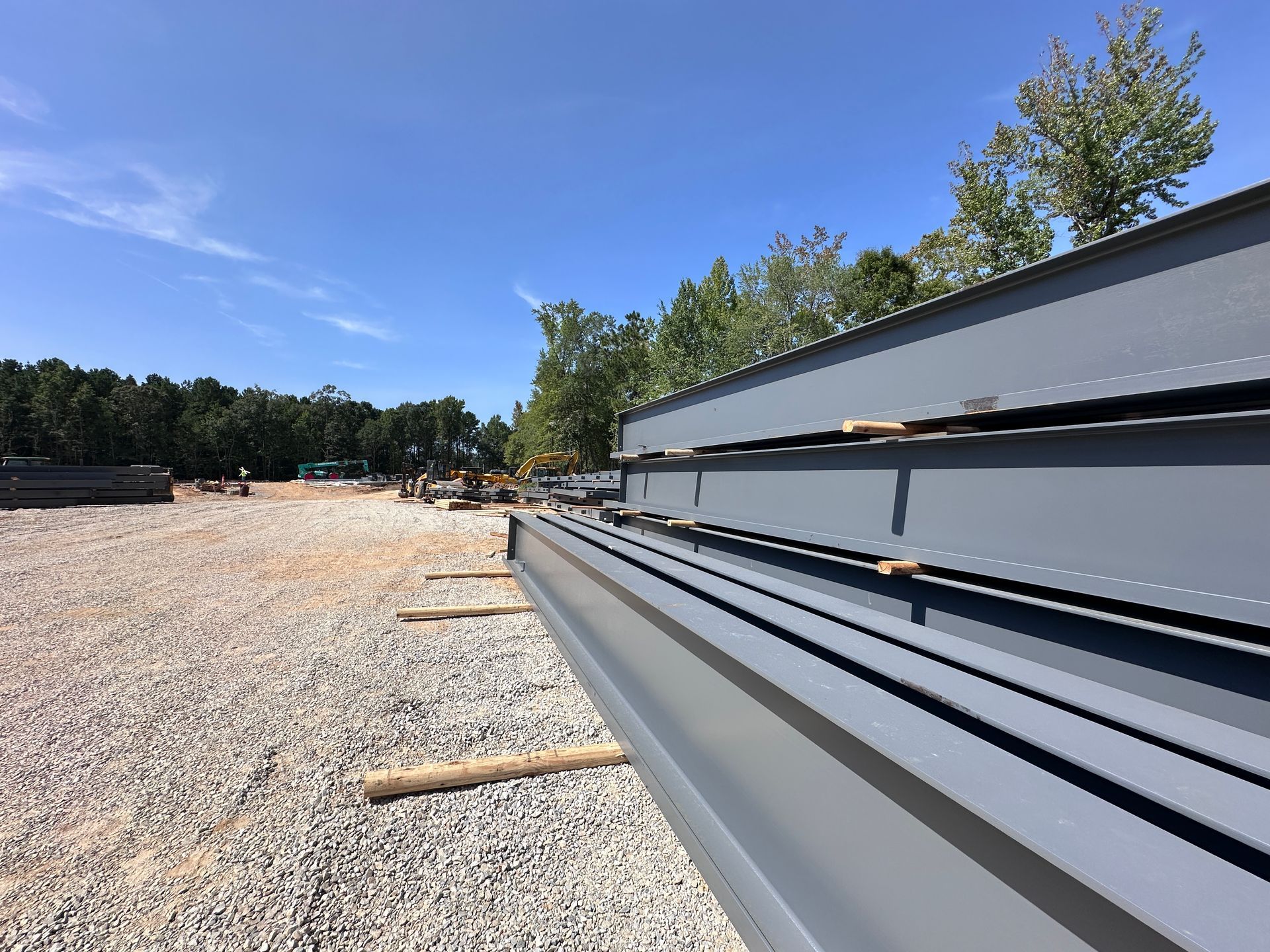 Stacked gray metal beams on a gravel lot under a clear blue sky