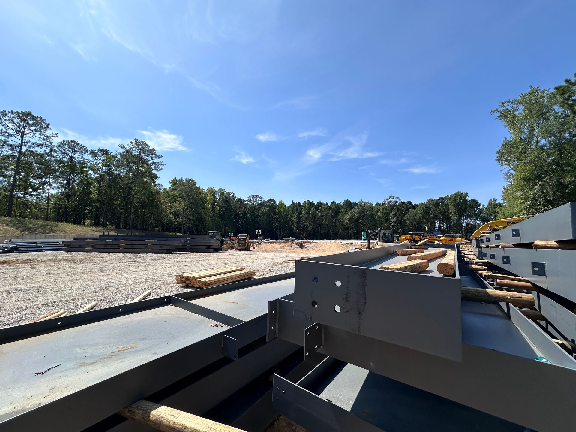 Construction site with steel beams in foreground under blue sky, surrounded by trees and gravel ground