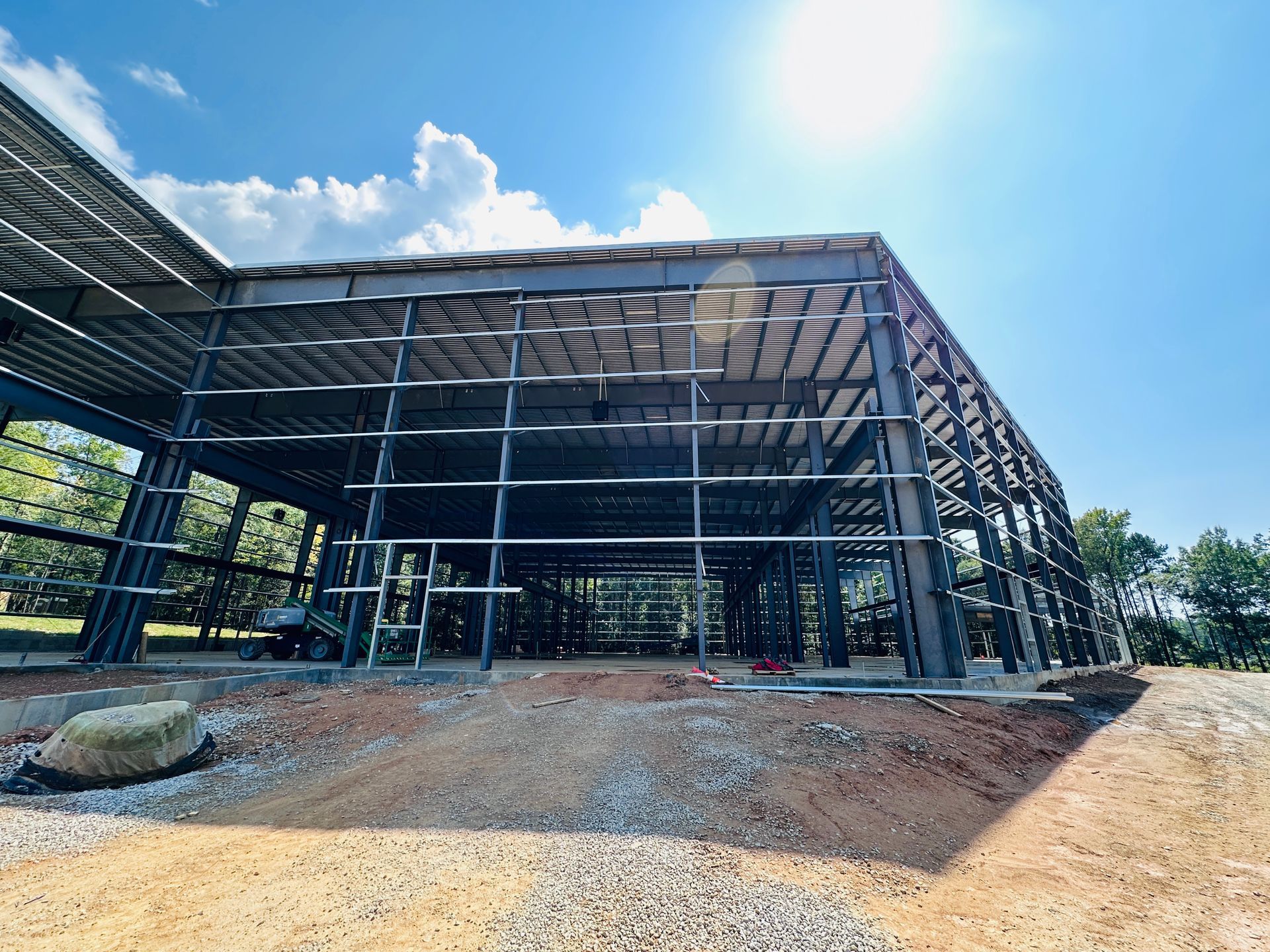 Large steel-frame building under construction with open sides and a dirt foreground under a blue sky
