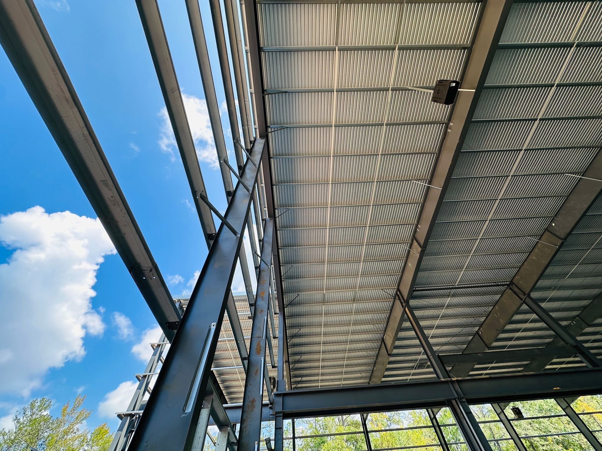 Steel roof beams and corrugated metal canopy against a blue sky with clouds