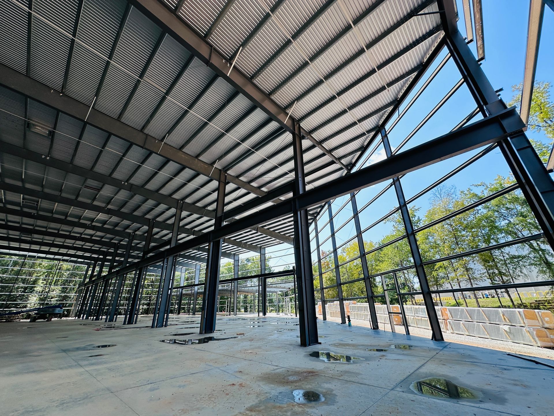 Open-air steel pavilion with a corrugated metal roof, concrete floor, and trees visible through the sides.