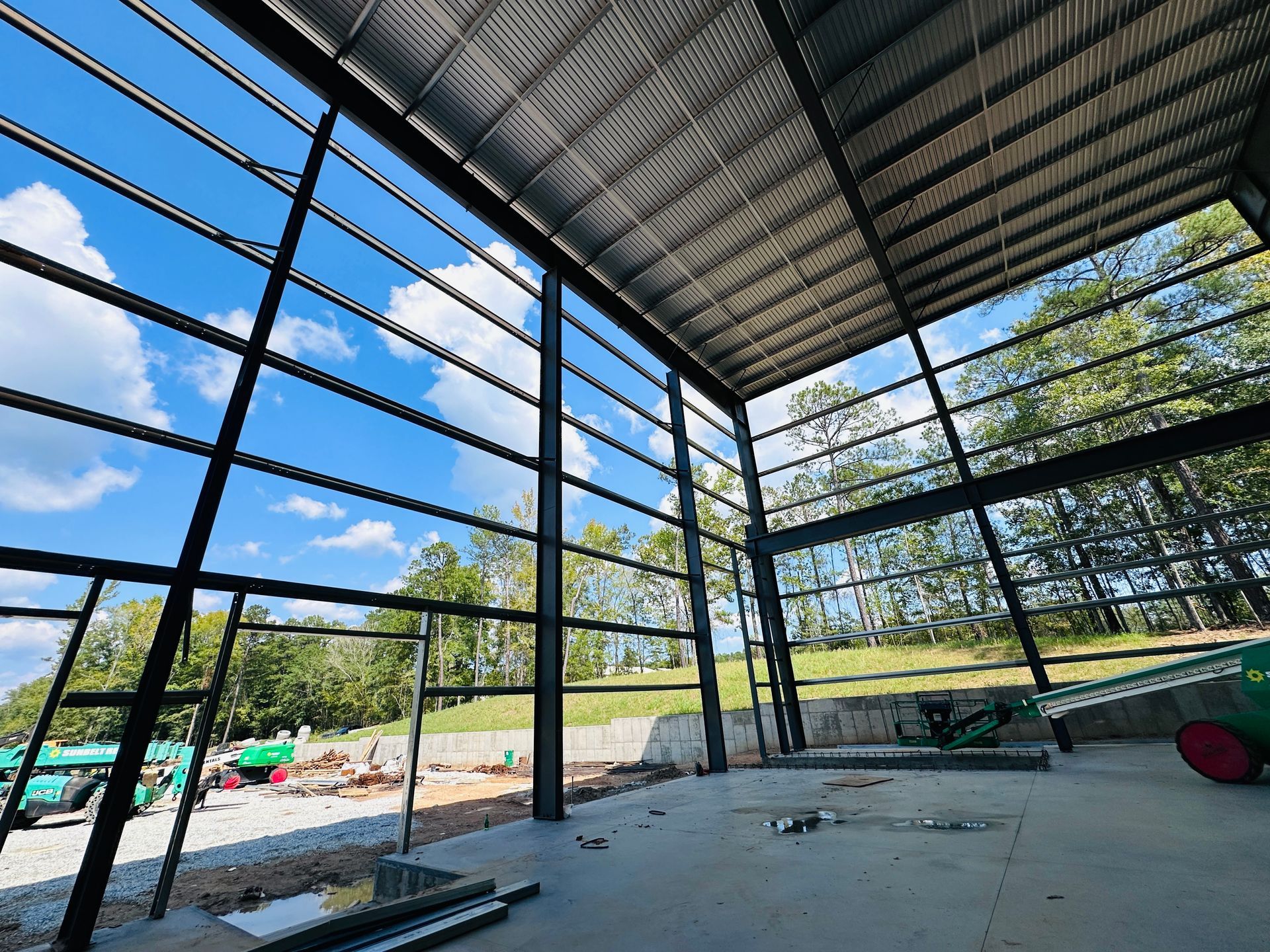 Open-air pavilion with steel beams, concrete floor, and a view of trees and sky outside
