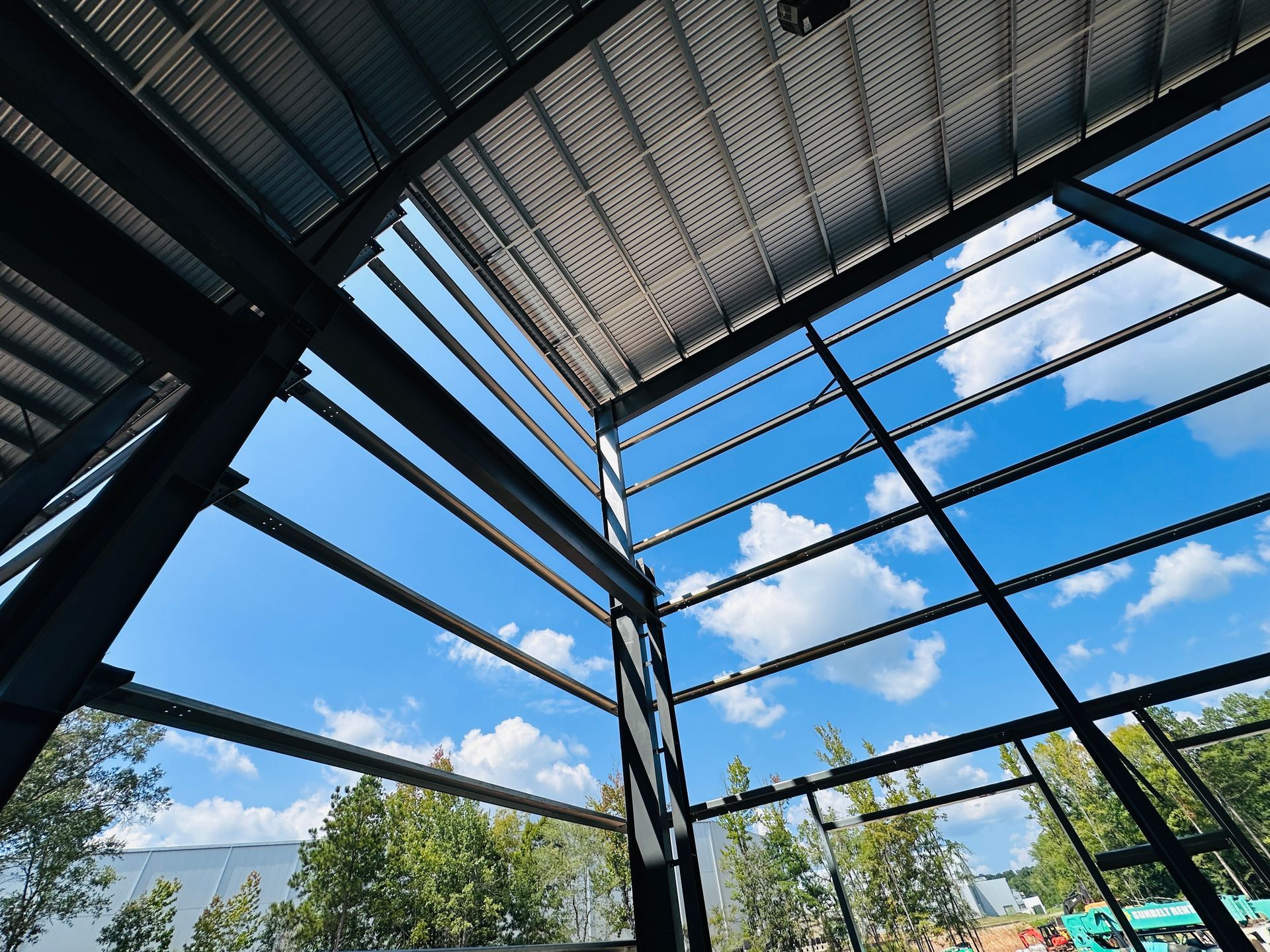 Metal roof structure with black beams framing a bright blue sky, clouds, and trees below