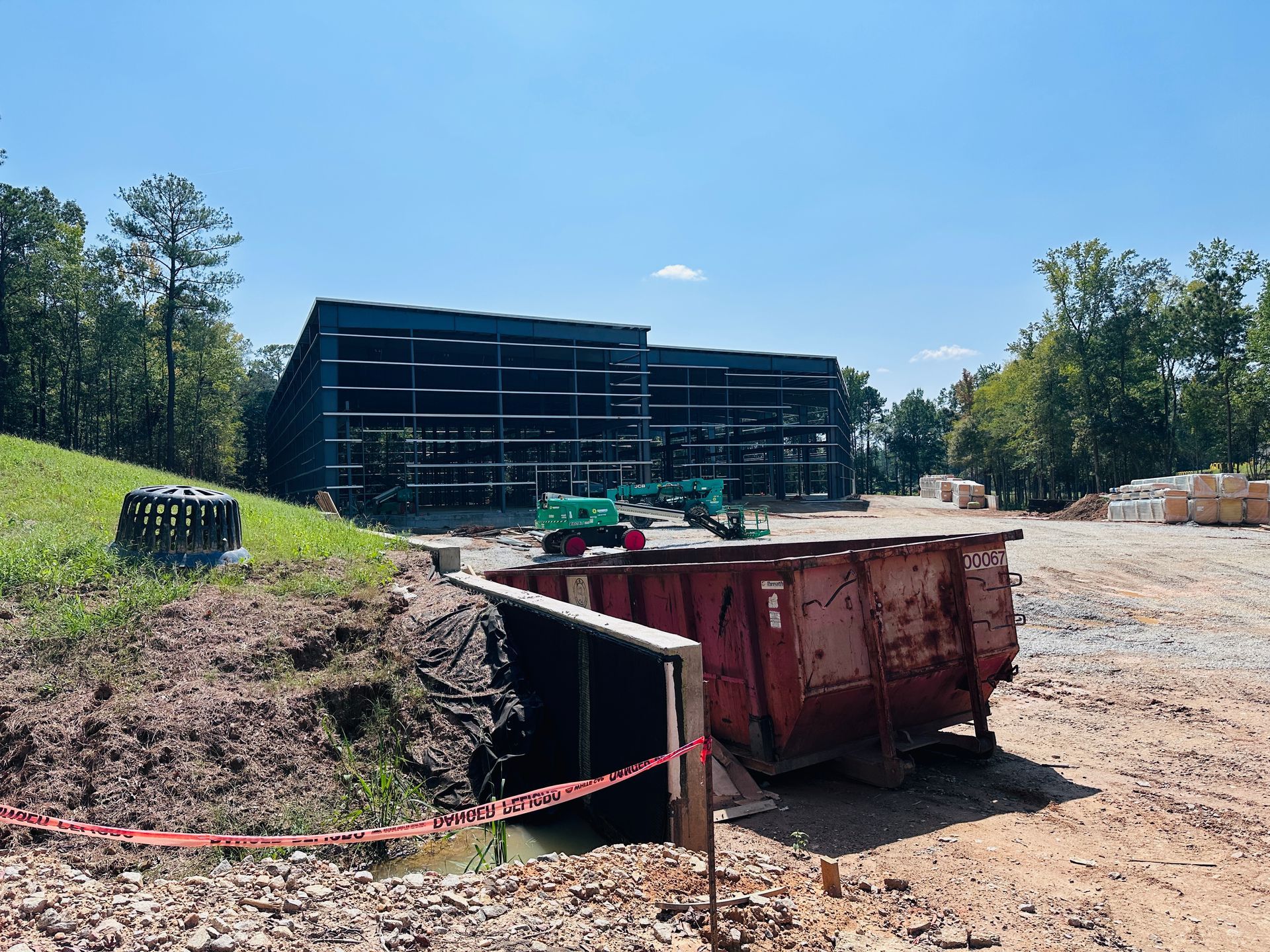 Construction site with deep trench, red dumpster, and large black structure under a blue sky.