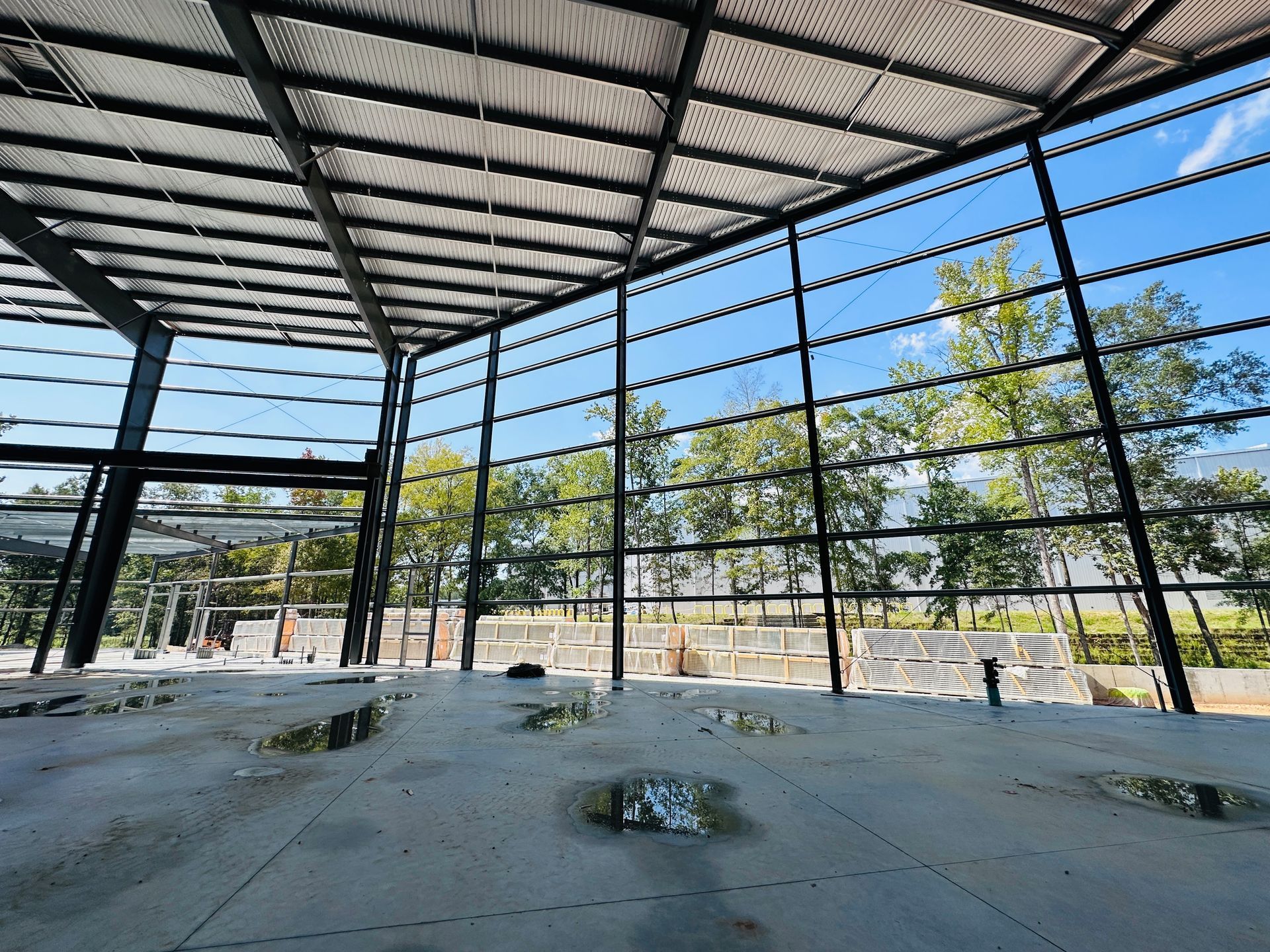 Empty metal-framed building interior with a concrete floor and open walls overlooking trees and blue sky