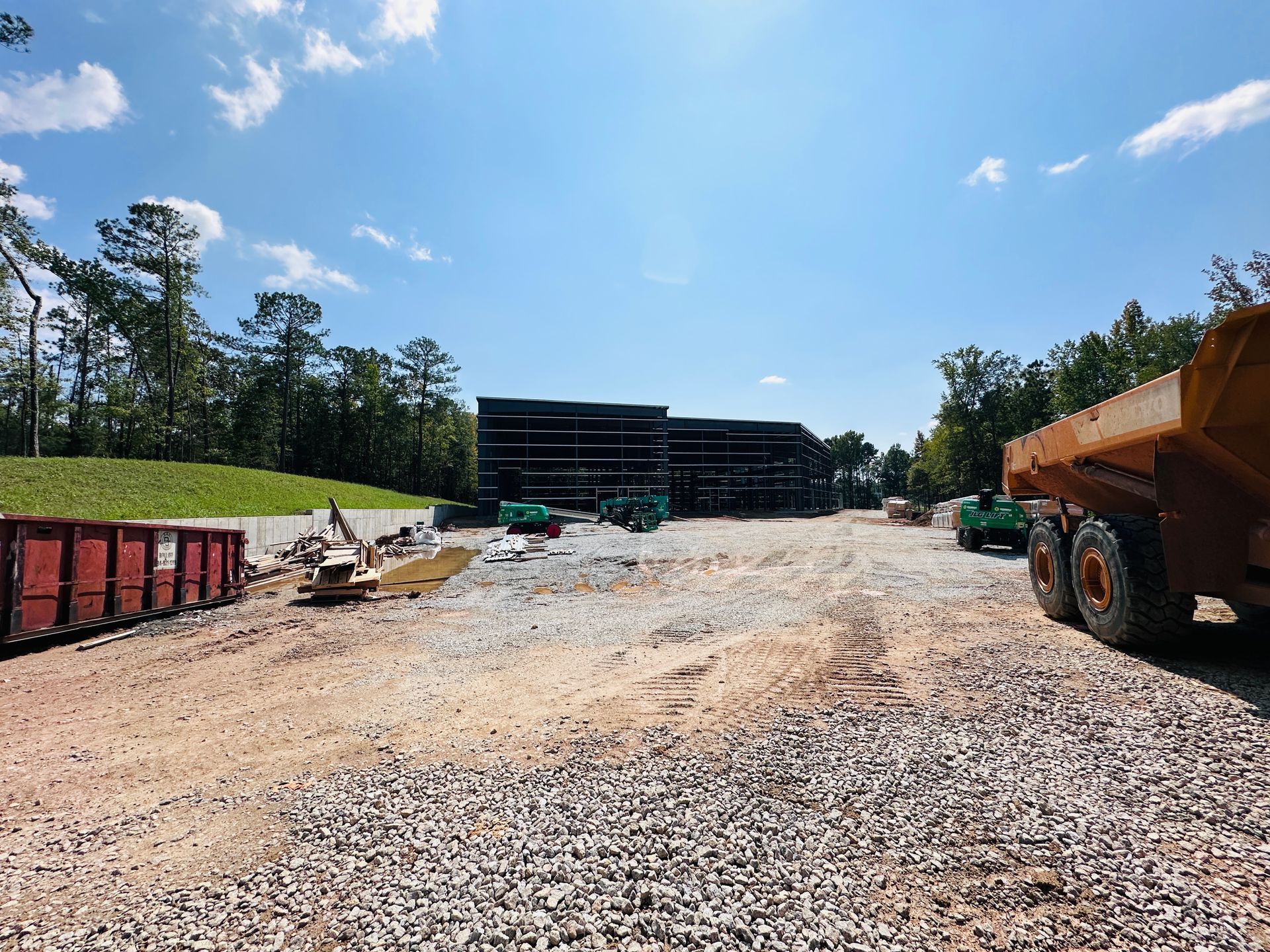 Gravel lot with construction materials, trees, and a large building under a bright blue sky