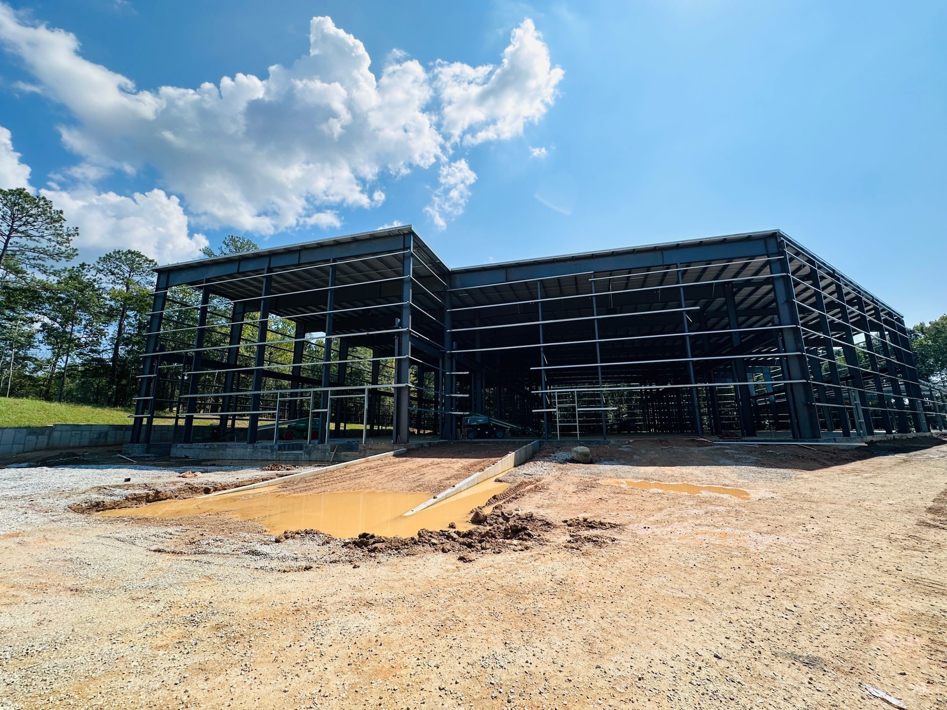 Partially built steel-frame building on a gravel lot under a blue sky with clouds