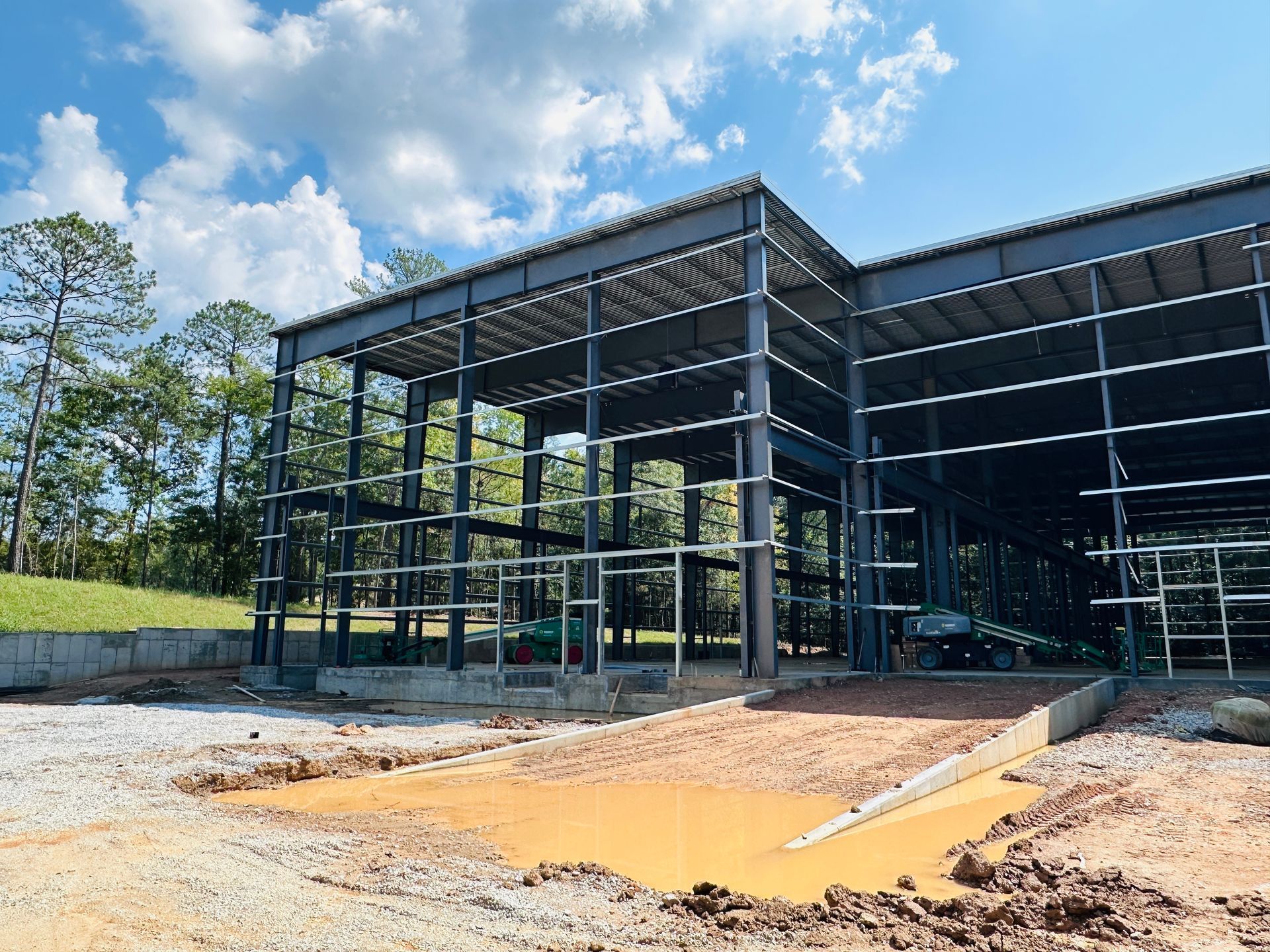 Steel-framed building under construction with exposed foundation and dirt foreground under a blue sky