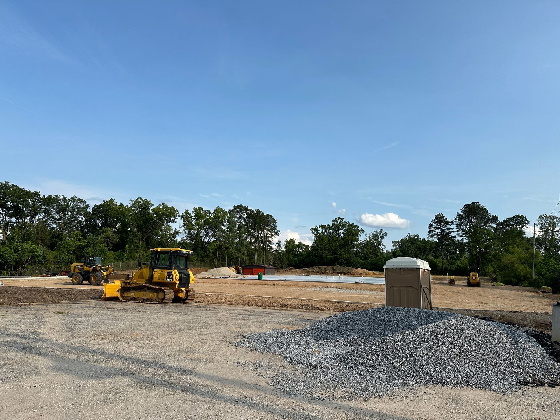 Construction site with a yellow bulldozer, gravel pile, and portable toilet under a clear blue sky.