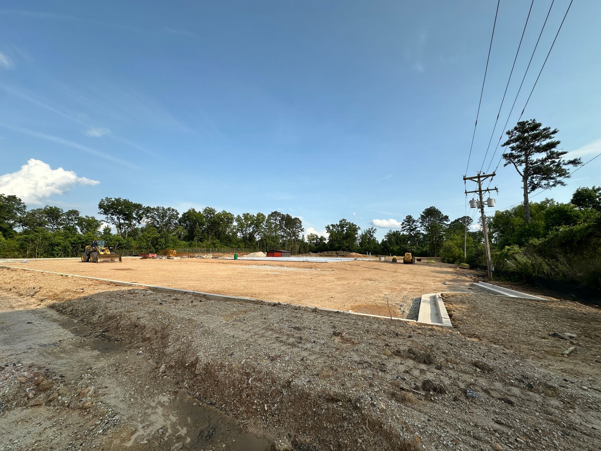 Bare dirt construction site with gravel foreground, trees in background, and power lines under a blue sky
