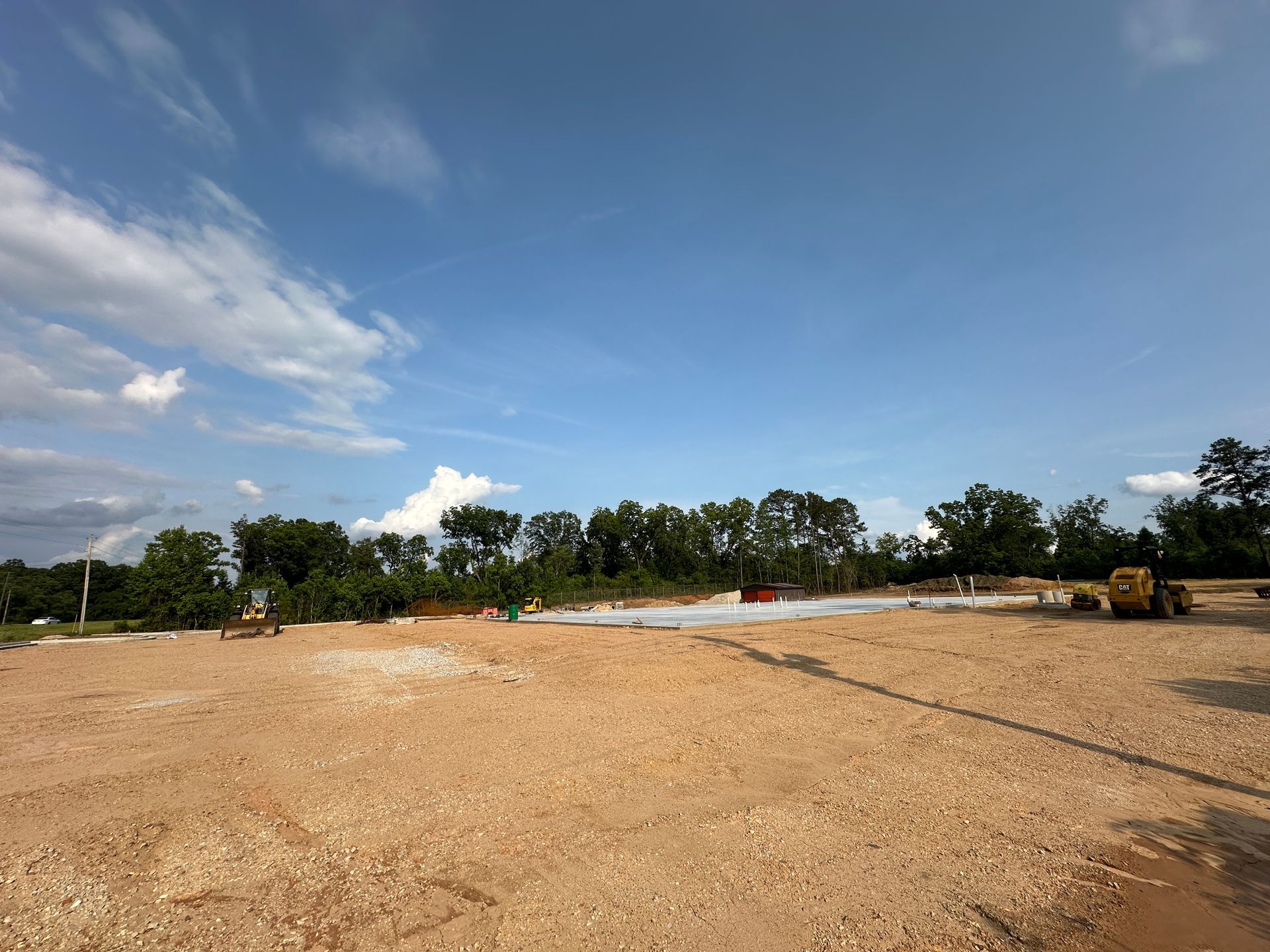 Wide dirt lot under blue sky, with trees in the distance and construction vehicles on the right.