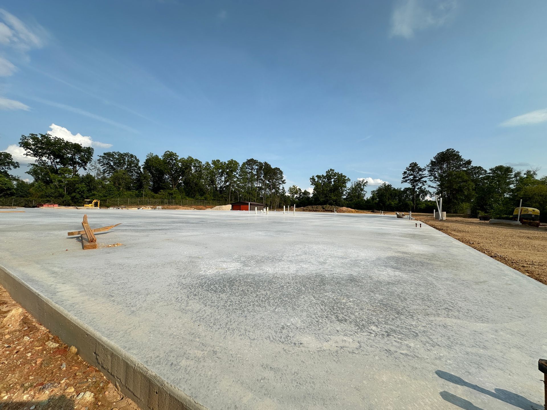 Concrete foundation slab at a construction site under a blue sky, with trees and dirt around it