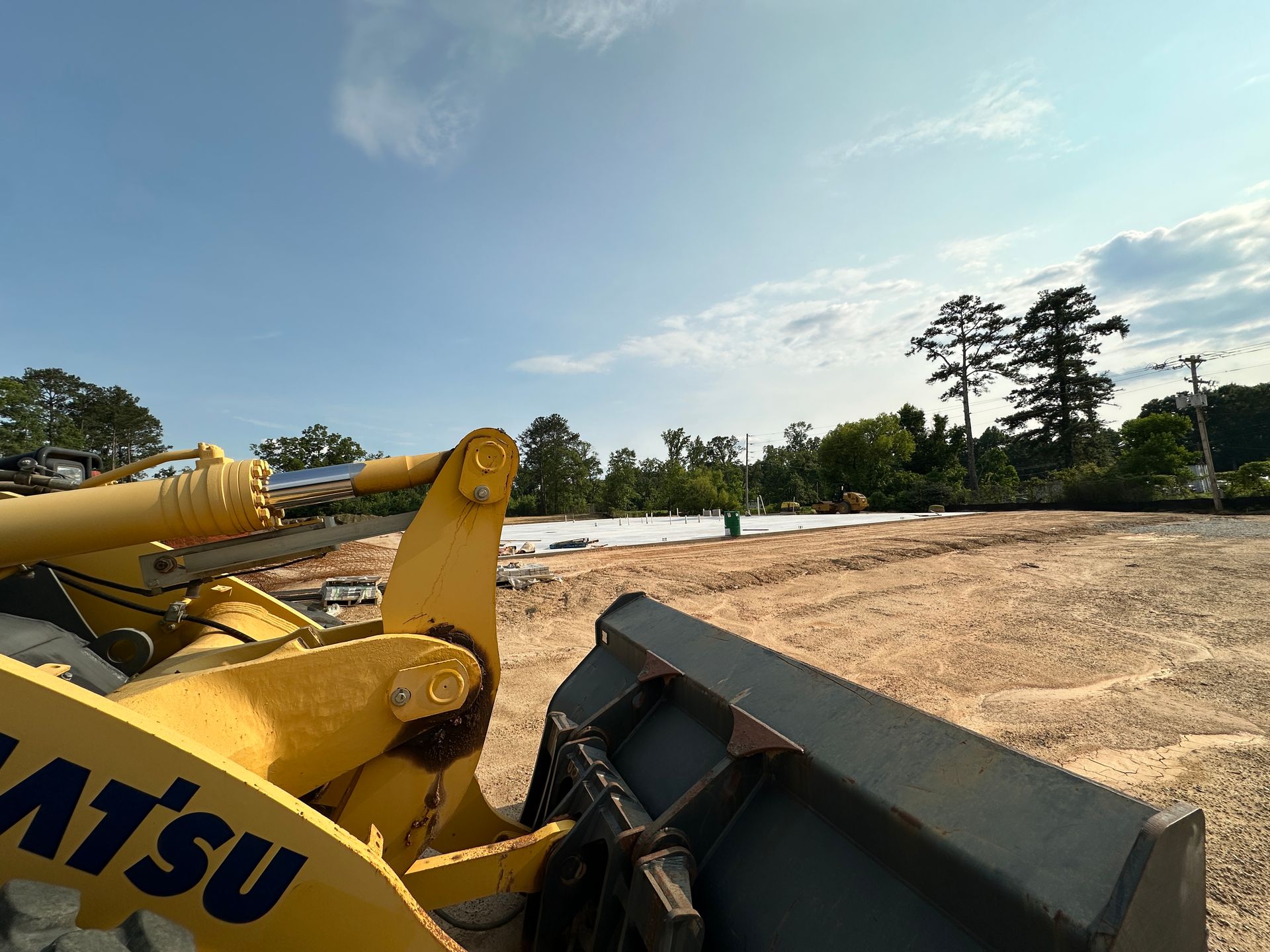 Construction site with dirt piles, heavy equipment, and a tree line under a partly cloudy blue sky