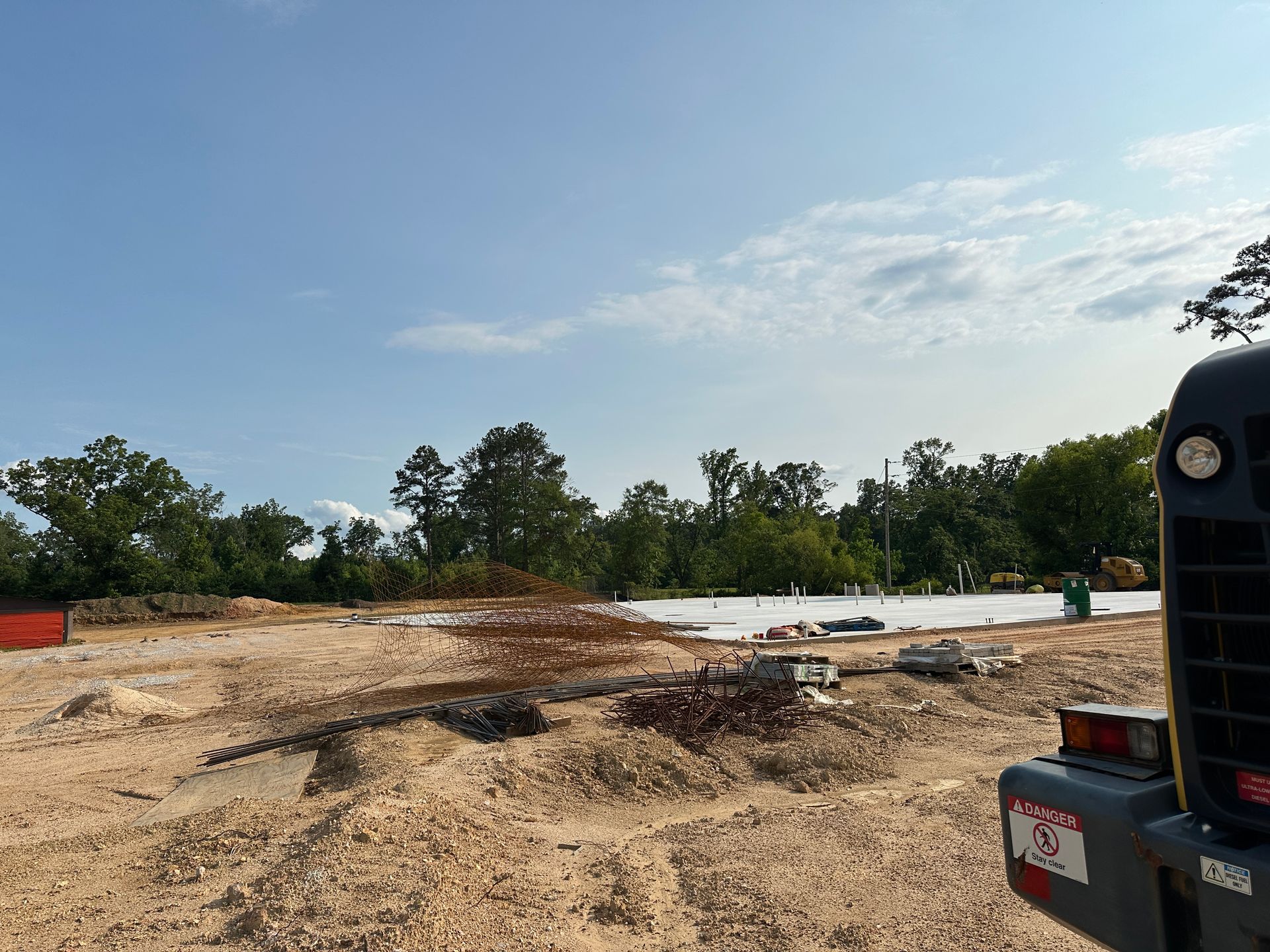 Cleared dirt lot with scattered debris, trees in the background, and a vehicle at right under a blue sky