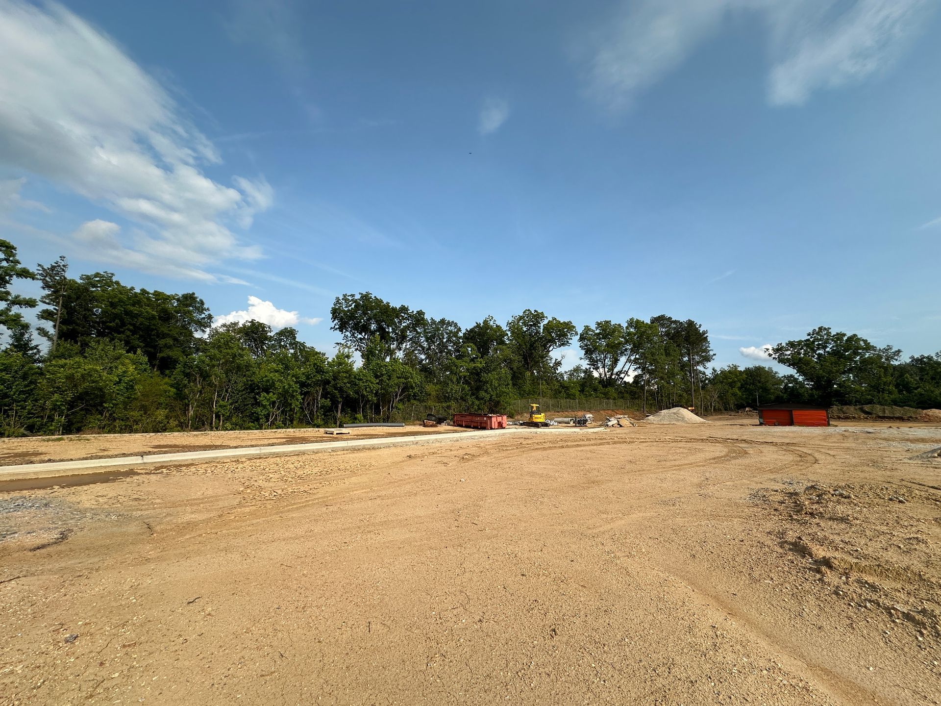Wide dirt road under a blue sky, bordered by green trees and a few distant vehicles.