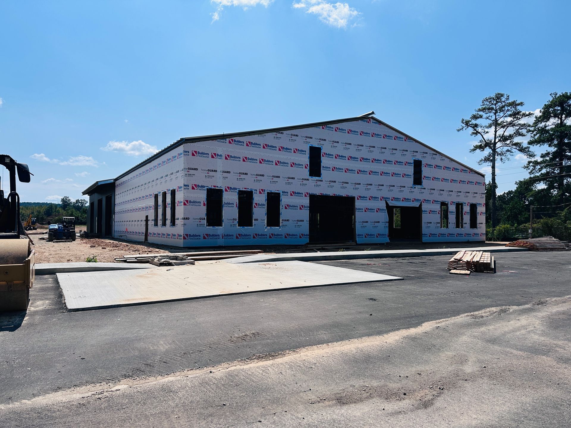 Construction site with dirt piles, heavy equipment, and a tree line under a partly cloudy blue sky