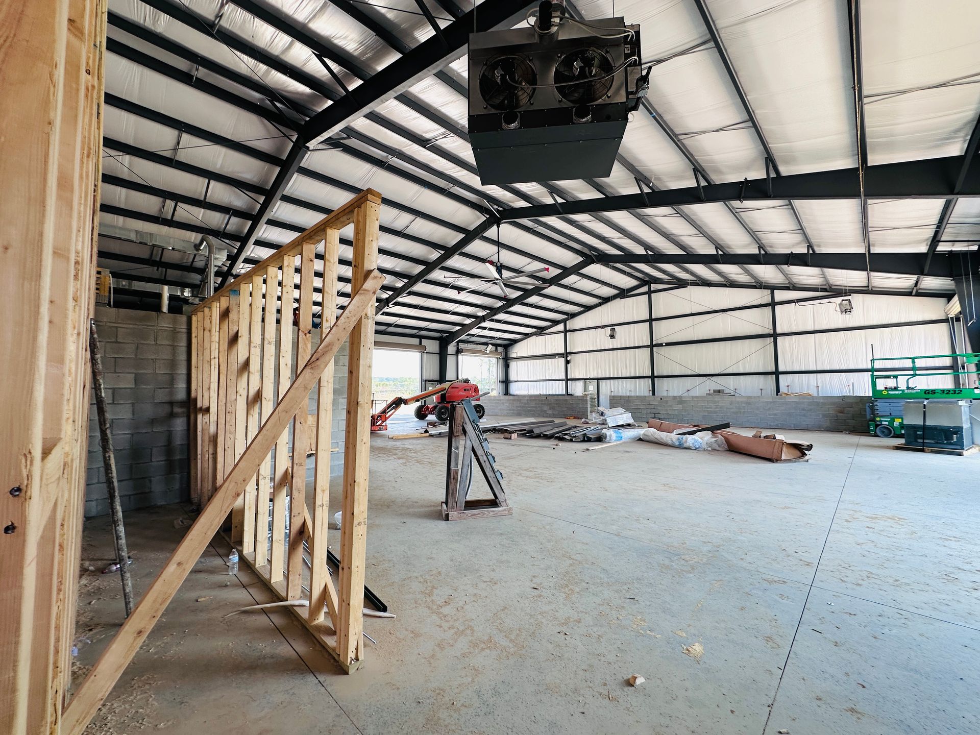 Interior of an unfinished warehouse with exposed framing, metal roof trusses, and scattered construction materials.