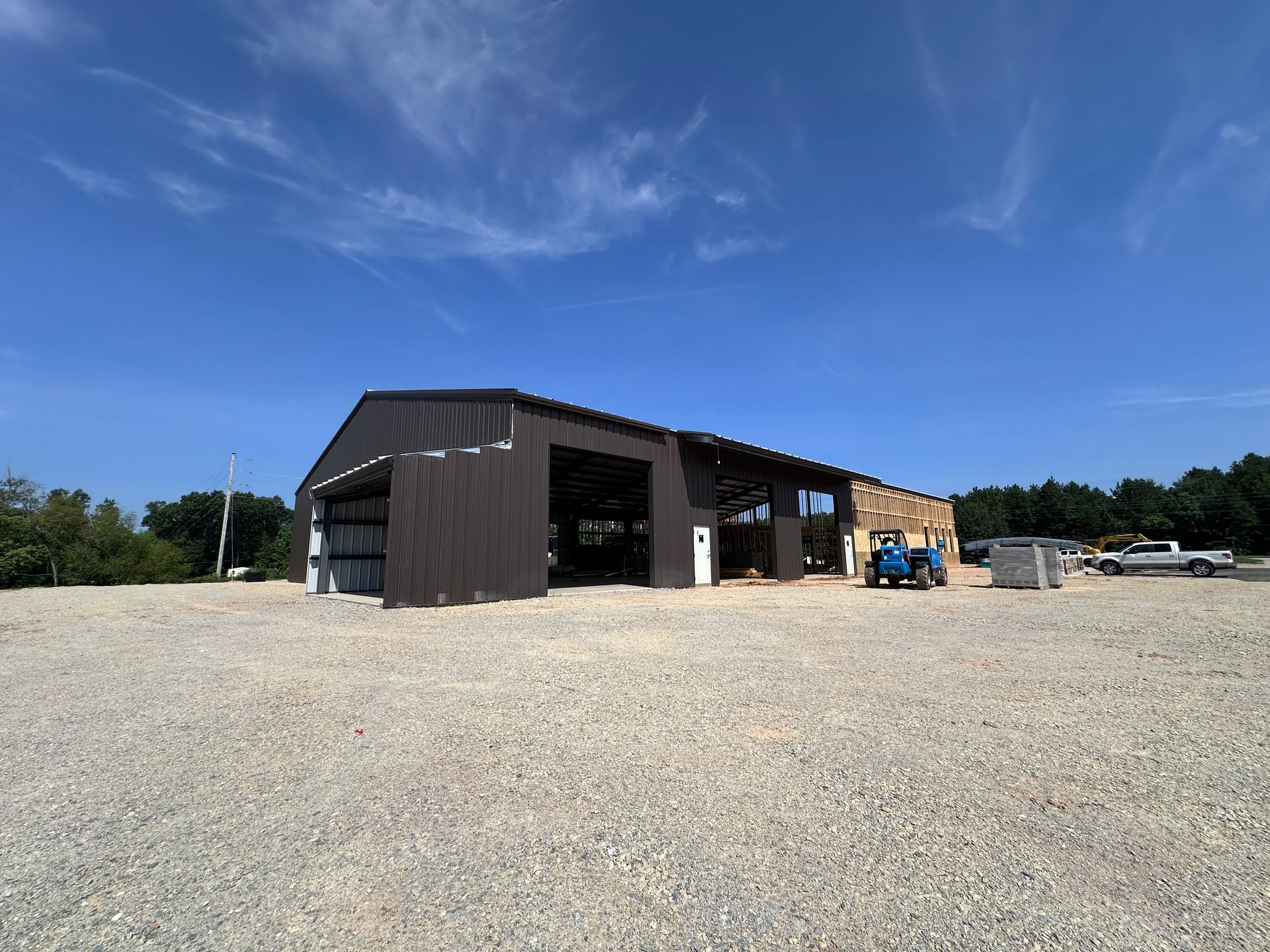 Large dark barn-like building on a gravel lot under a clear blue sky, with a small blue tractor nearby.