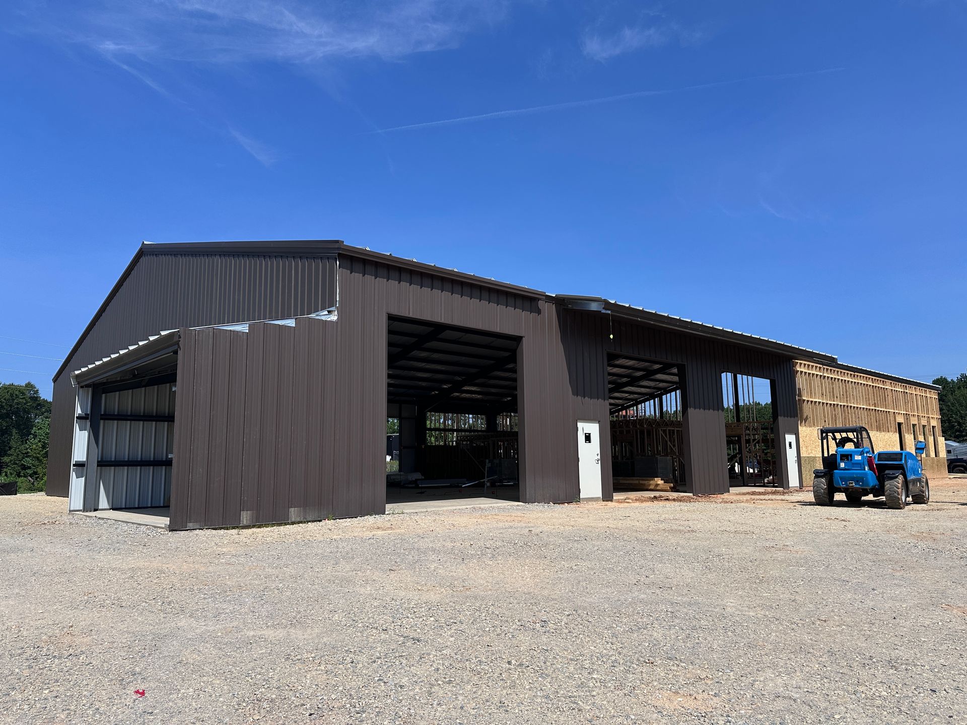 Large metal barn with open bays on a gravel lot, with a blue utility vehicle parked beside it.