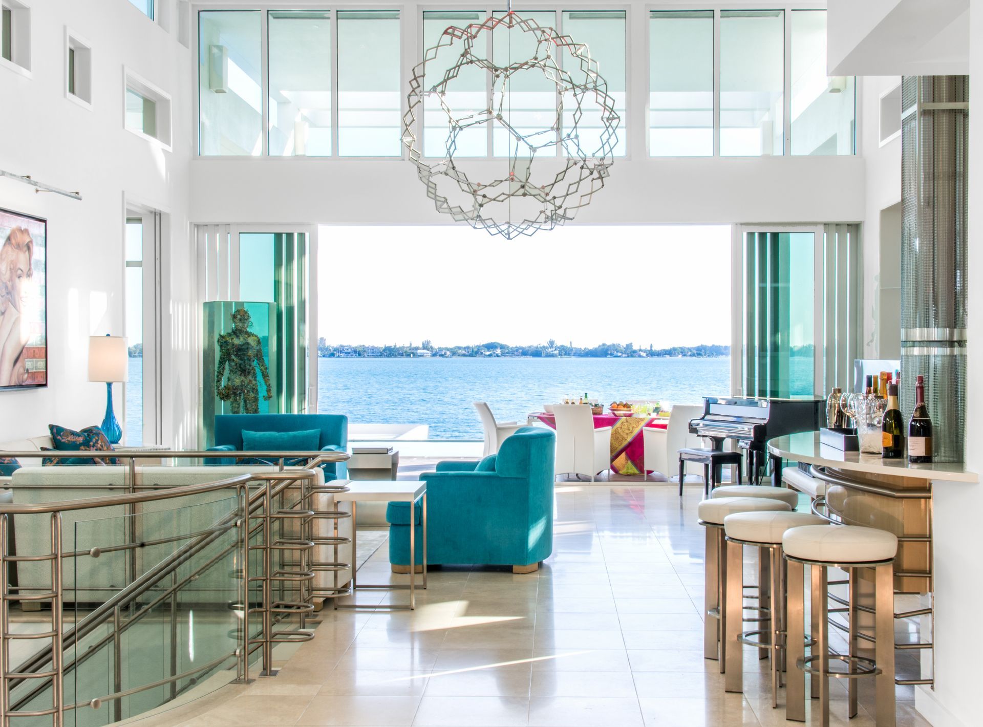 Spacious, modern interior with blue accents, expansive waterfront view. A chandelier hangs over a bar with stools.