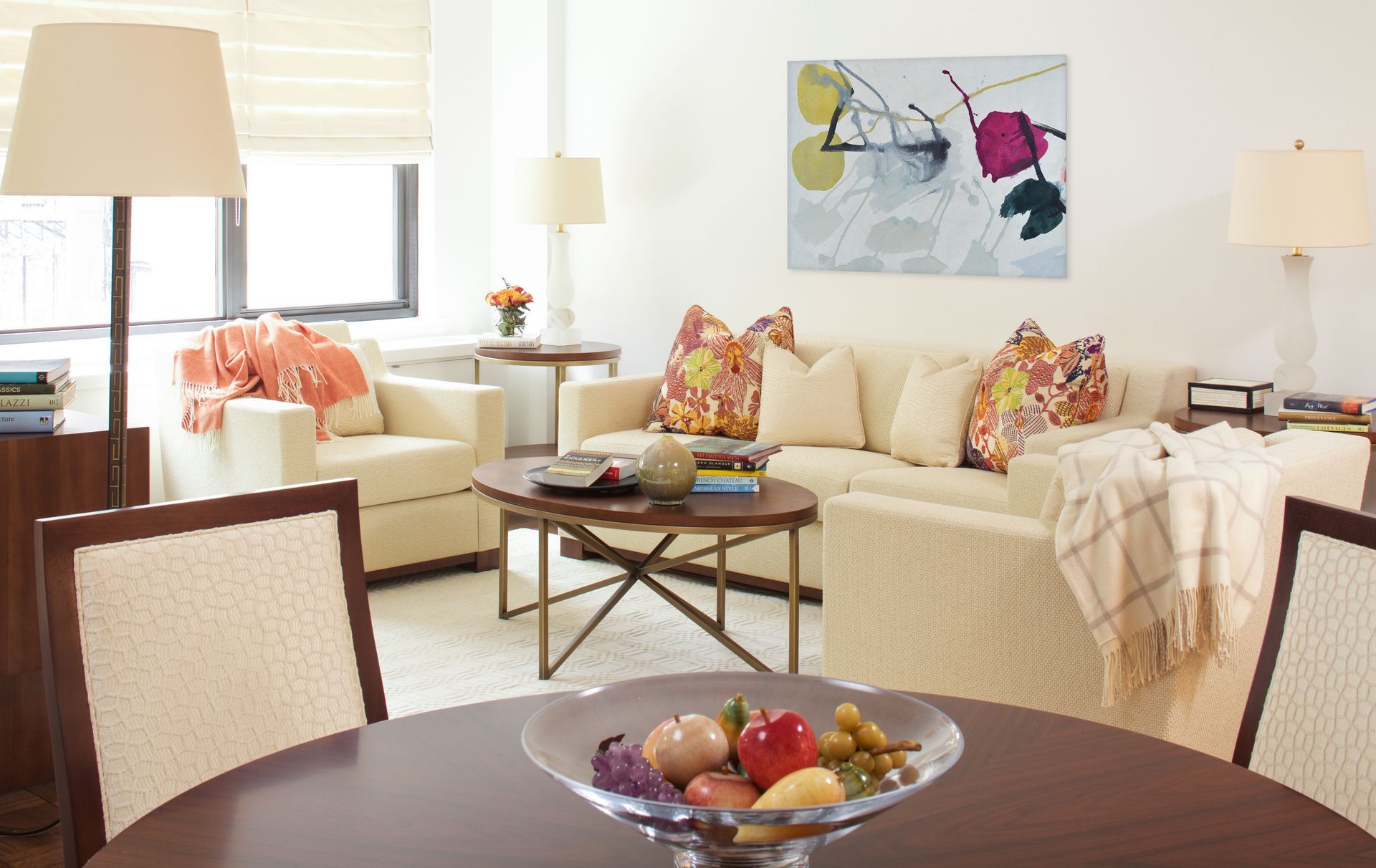 Living room: white sofa, armchairs, coffee table, dining table, and colorful floral pillows, painting on a white wall.