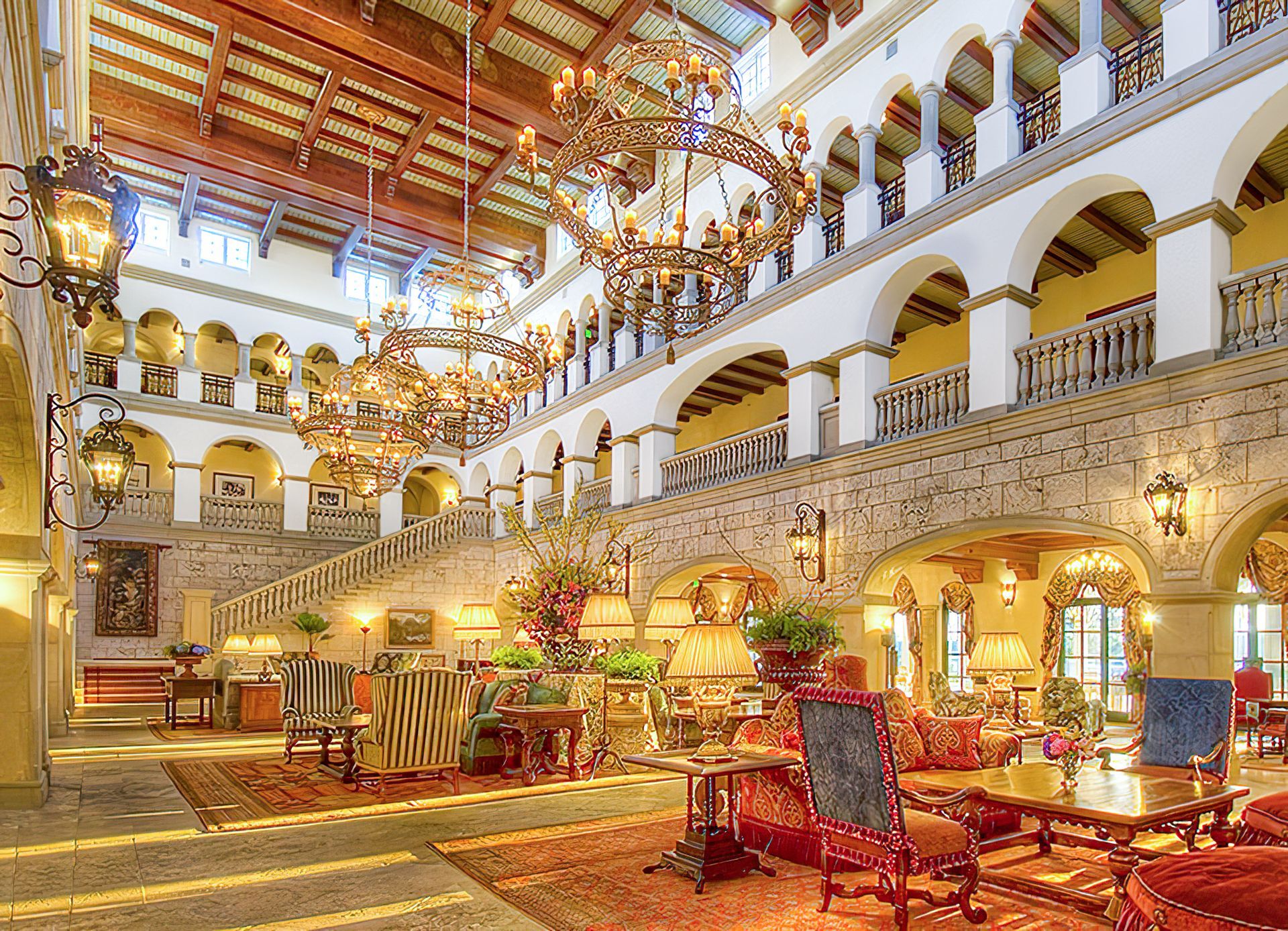 Grand hotel lobby with balconies, stone walls, chandeliers, and seating areas.