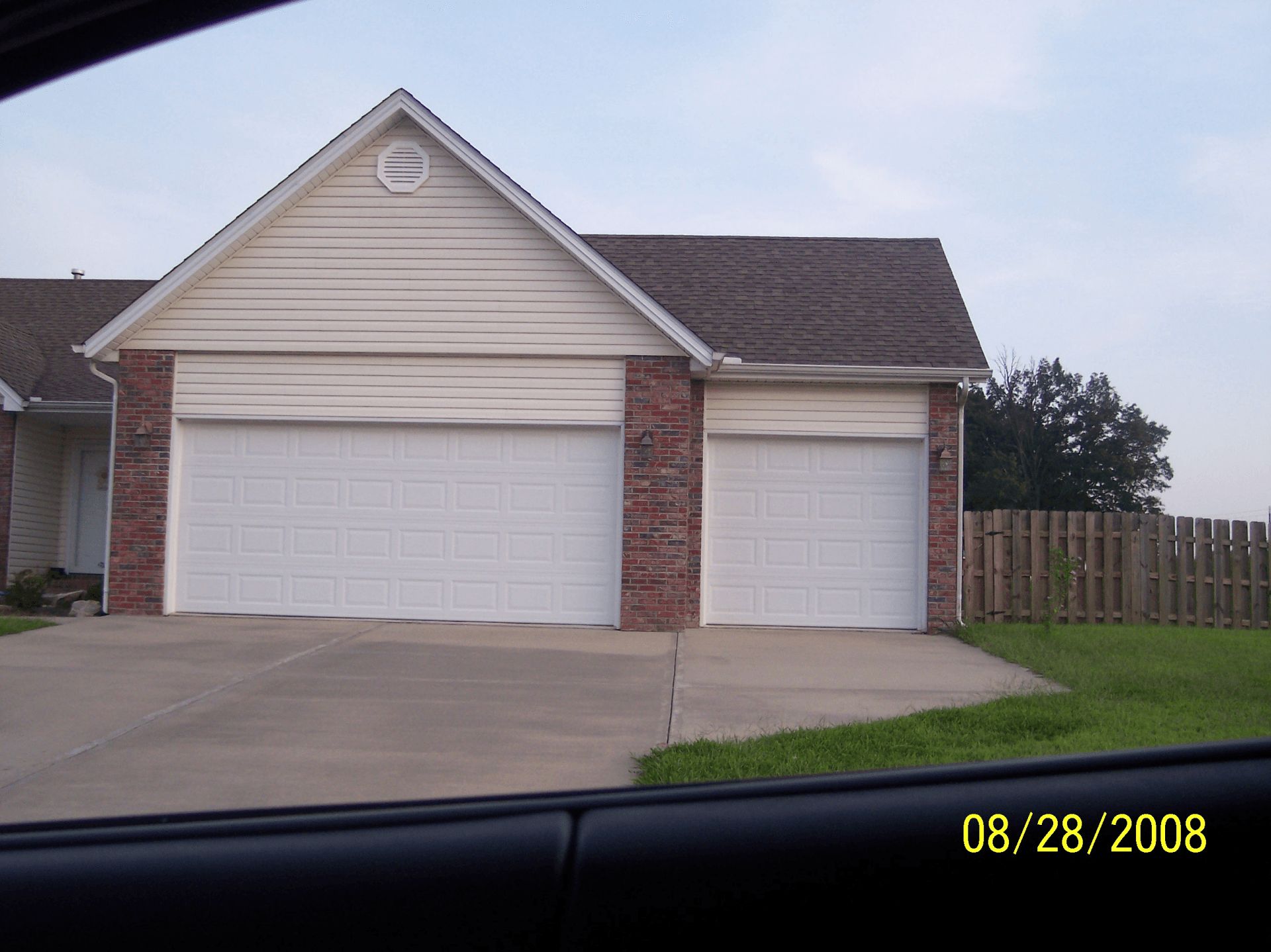 Suburban house exterior with two white garage doors, brick trim, driveway, and fenced yard.