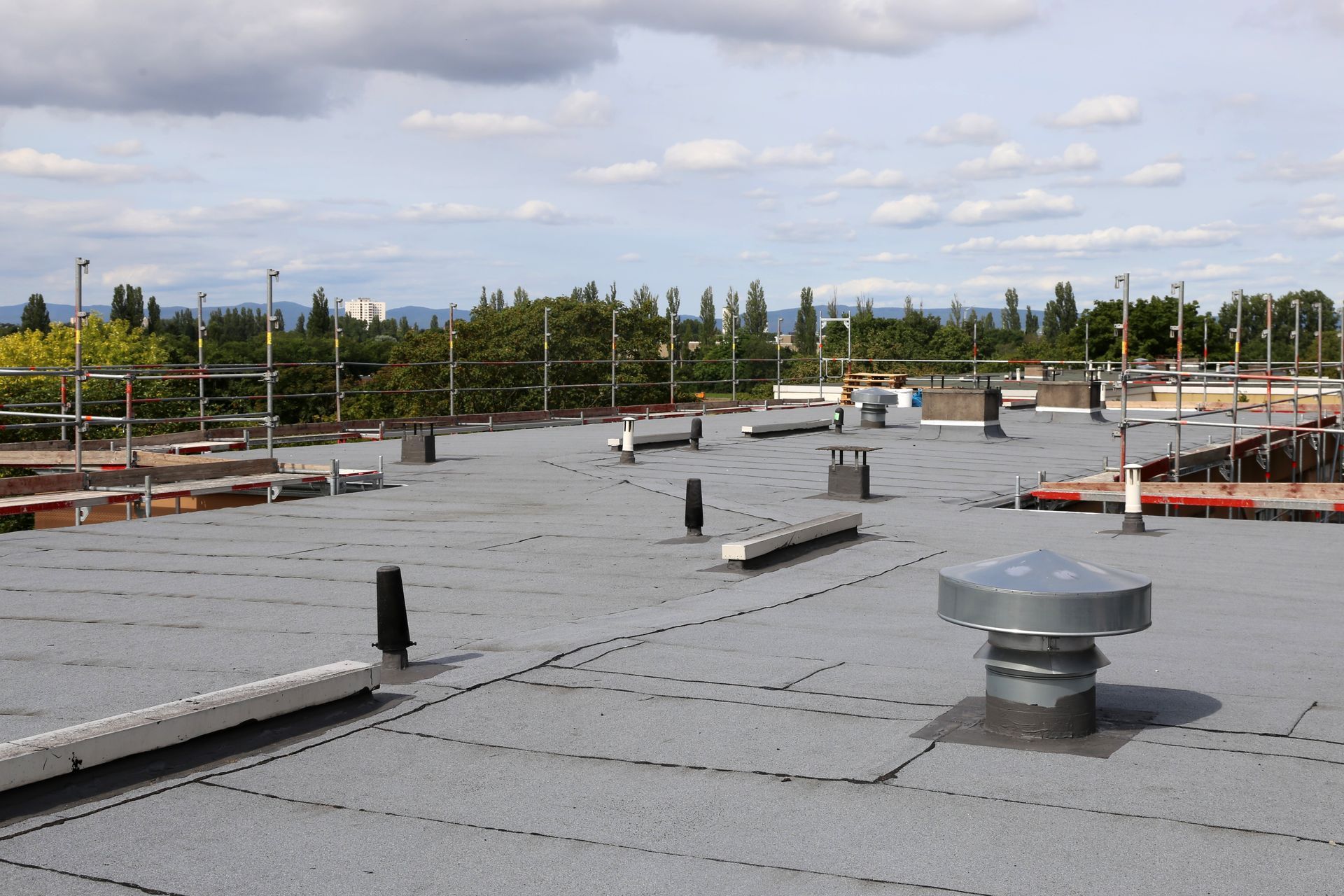 A flat commercial roof with grey membrane, ventilation pipes, and safety scaffolding against a cloudy blue sky.