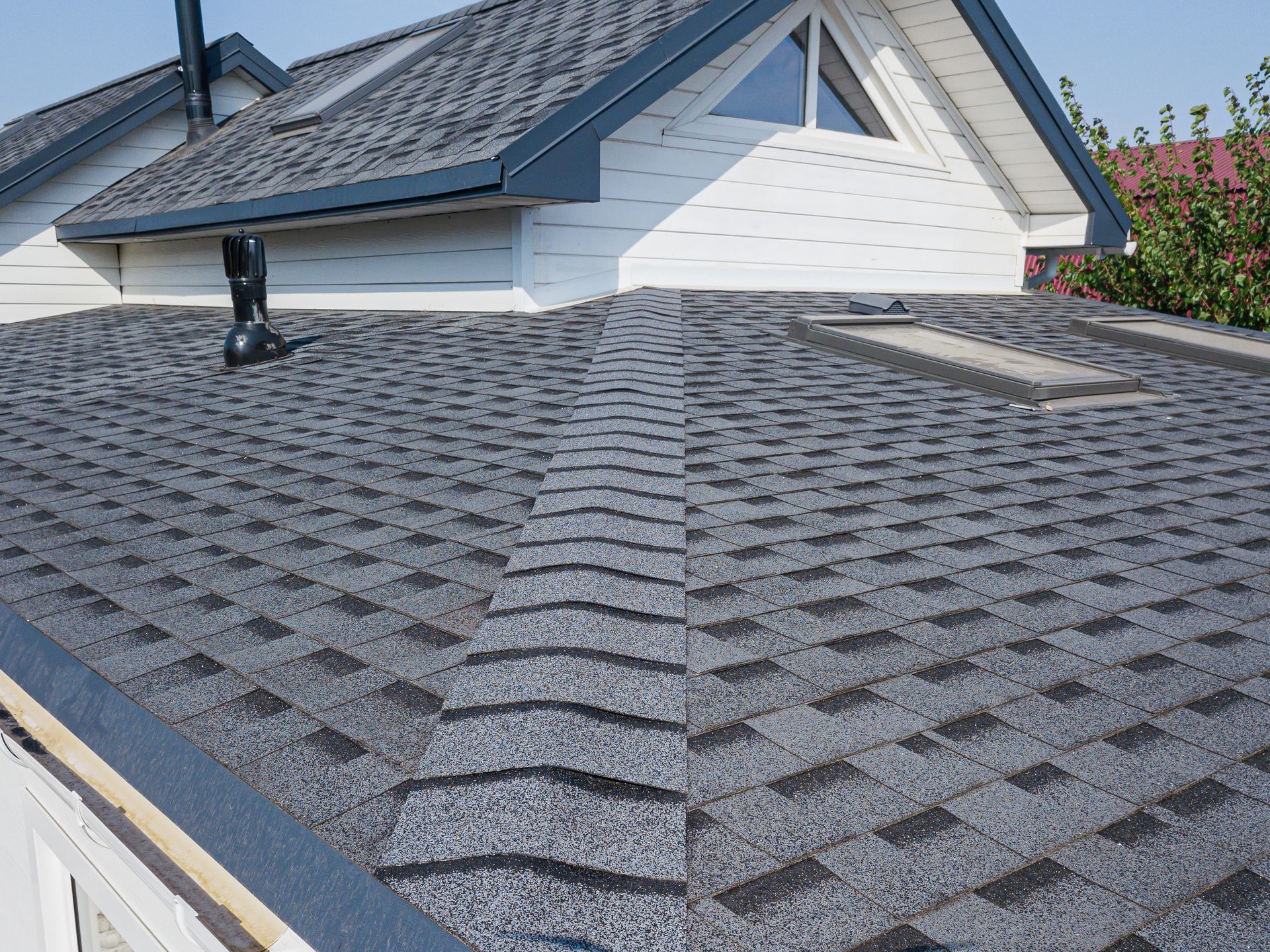 Close-up view of a residential shingled roof with a gabled upper section, light-colored siding, and skylights.