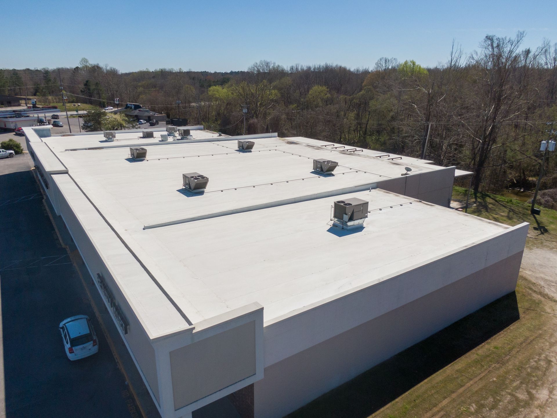 An aerial view of a flat, light-colored industrial building roof with several HVAC units, surrounded by trees and a road.