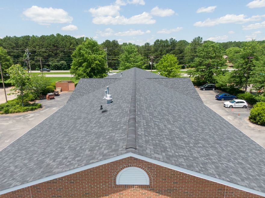 Aerial view of a gray shingled roof on a brick building with a parking lot and green trees in the background.