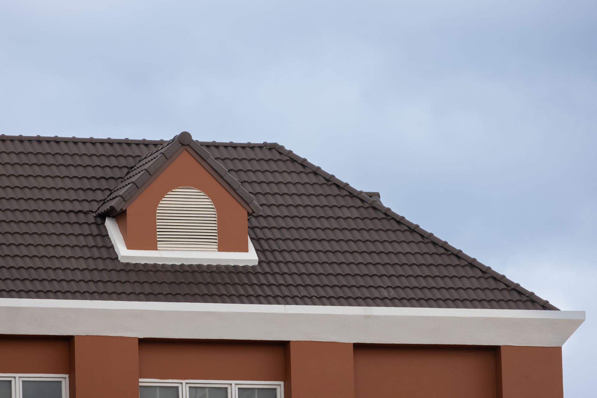 A brown tiled hip roof with a small, arched dormer window featuring white shutters, set against a cloudy sky.