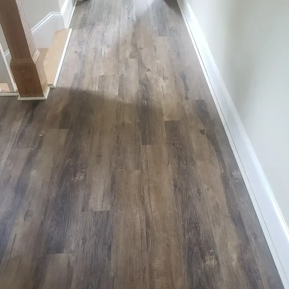 Wood-look flooring in a hallway with white trim and a staircase on the left. The floor has varying shades of brown.