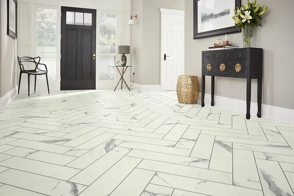 A brightly lit entryway with white marble-patterned floors laid in a herringbone pattern, black door, and gold accents.