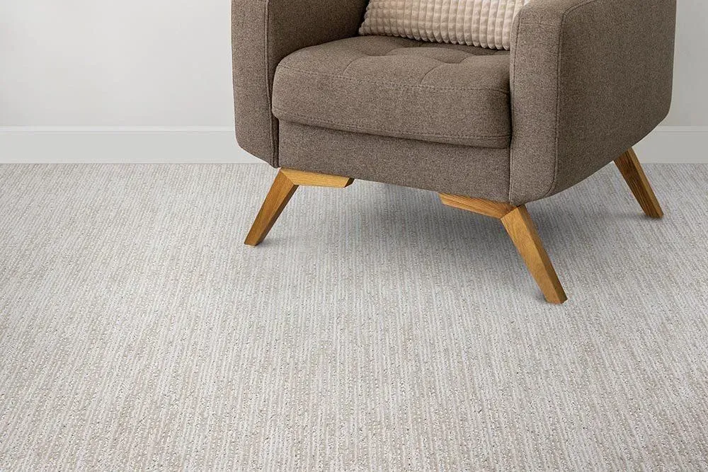 An armchair sits on a patterned beige carpet, against a white wall. The chair is brown, with light-colored wooden legs.