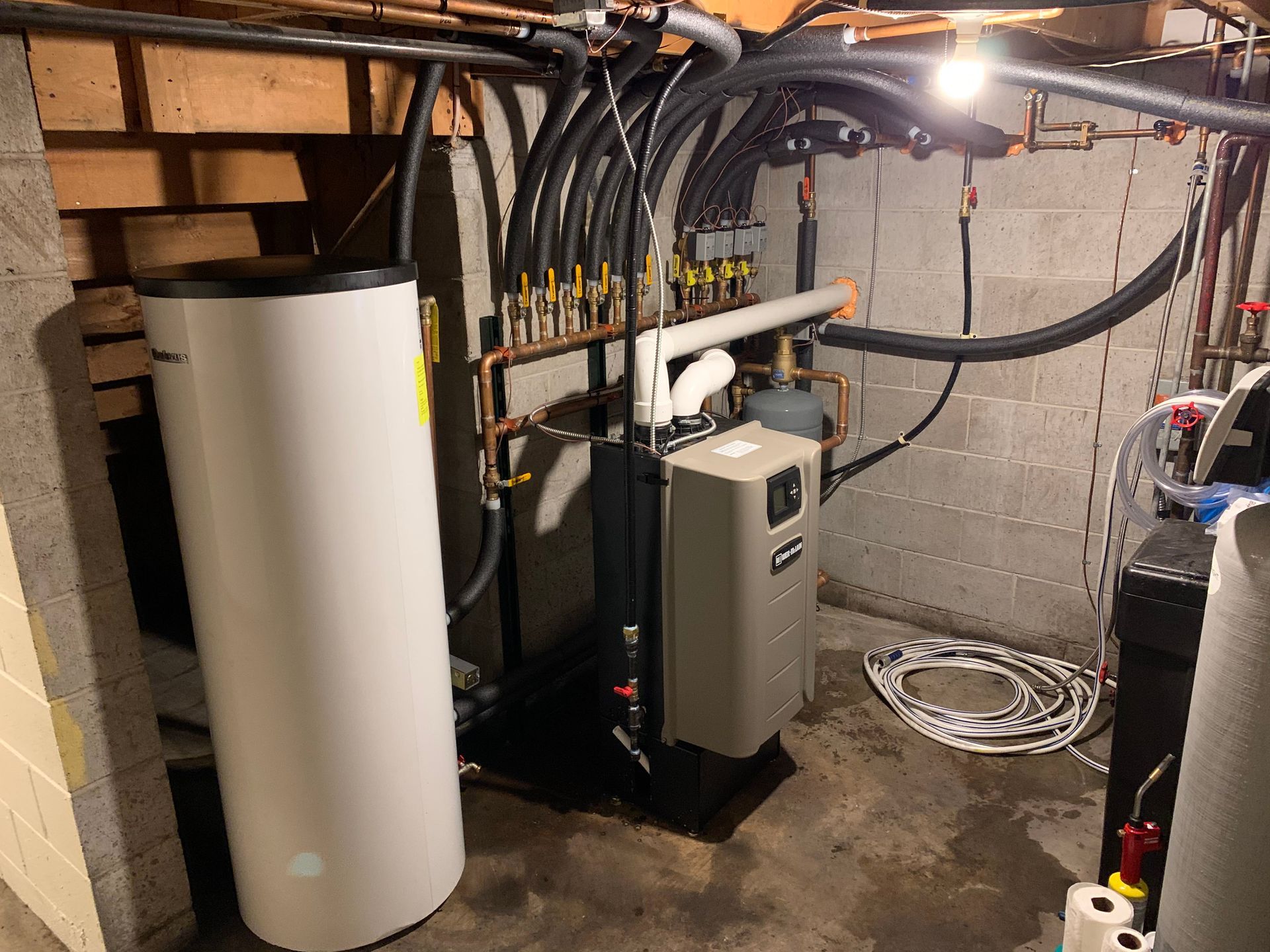 A white water heater is sitting in a basement surrounded by pipes.