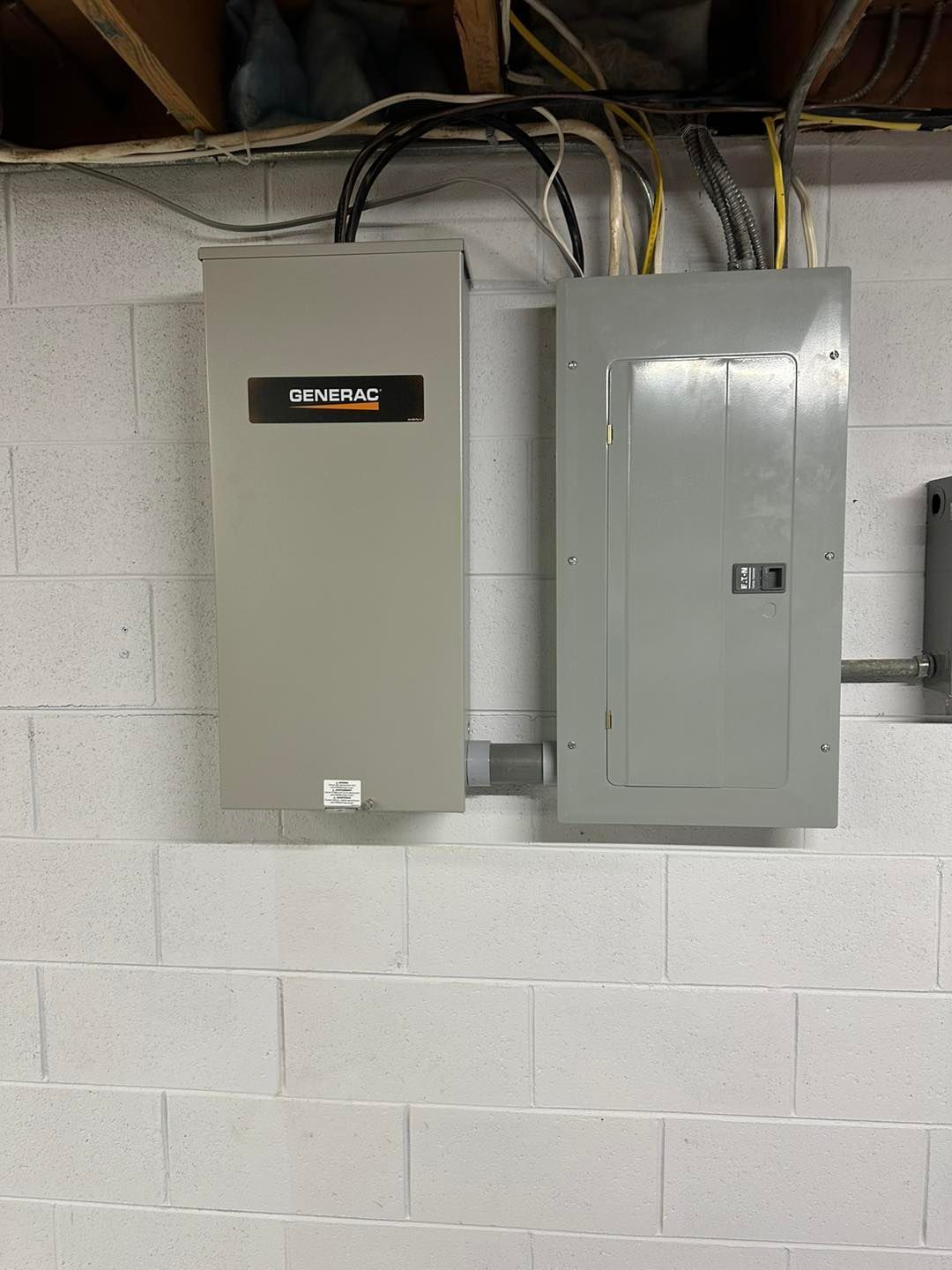 Two gray electrical panels mounted on a cinder block wall with exposed cables above.