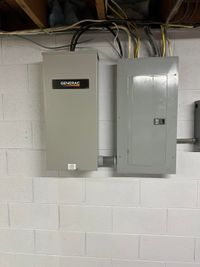 Two gray electrical panels mounted on a cinder block wall with exposed cables above.