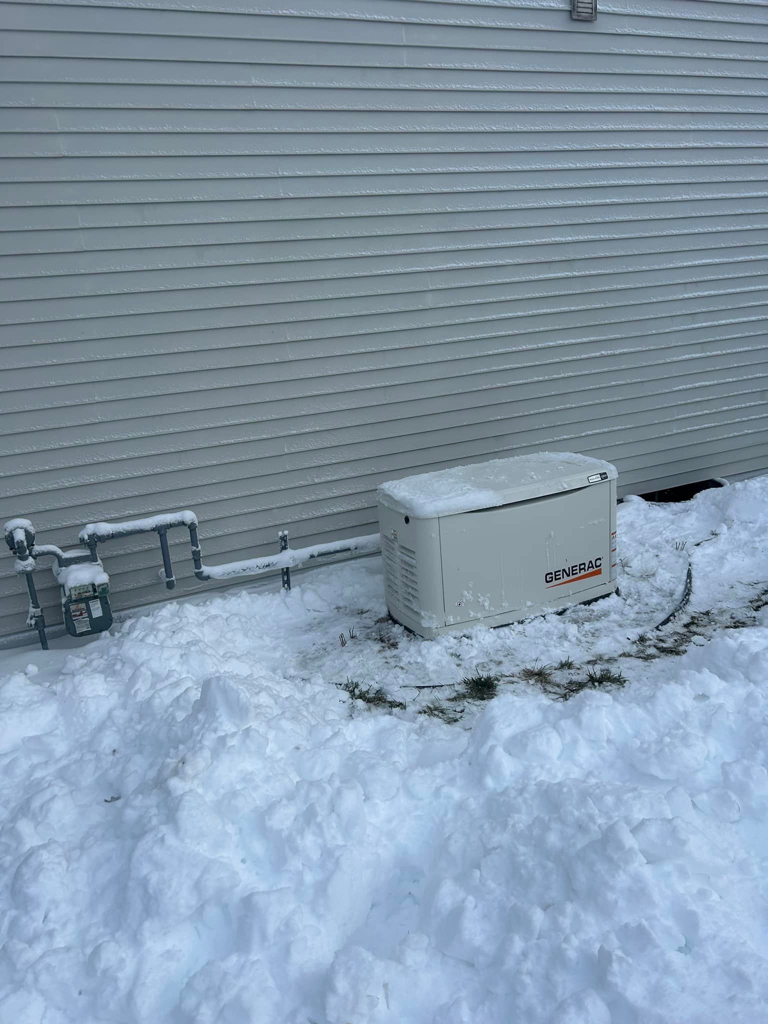 Snow-covered yard with a white outdoor unit beside a house wall and utility pipes.