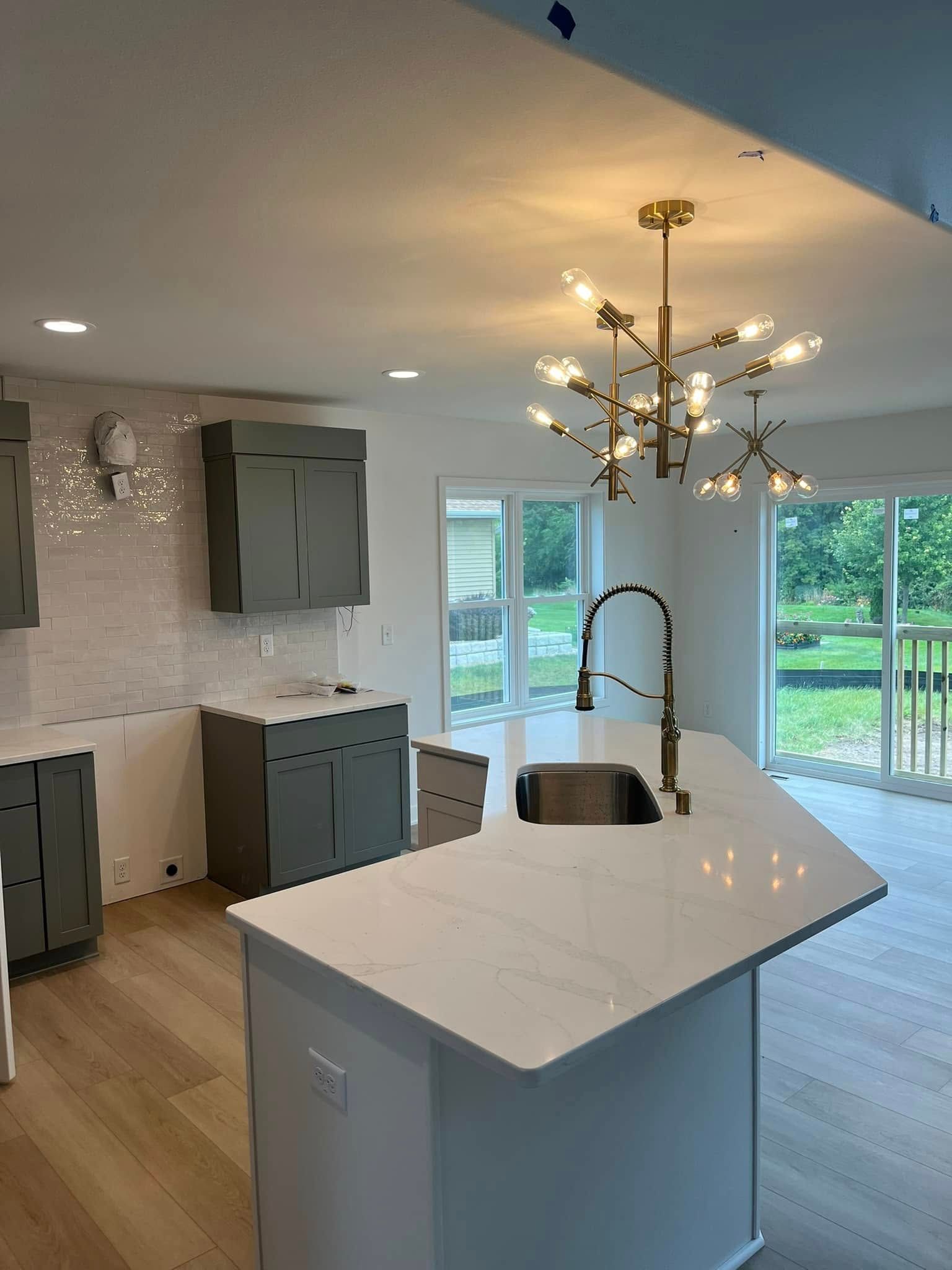 Modern kitchen with white island, gray cabinets, gold chandelier, and large sliding glass doors overlooking greenery