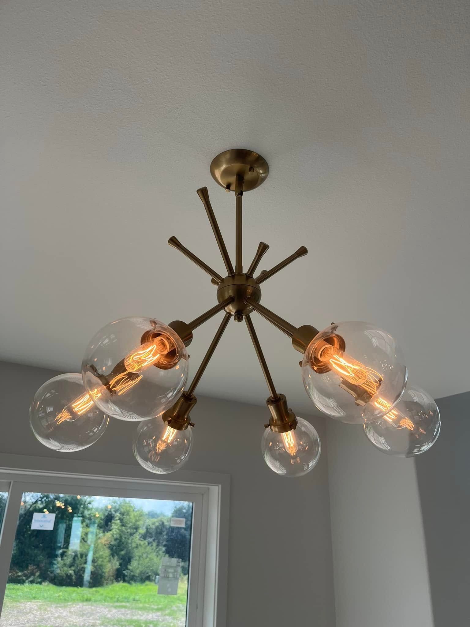 Modern brass starburst ceiling chandelier with clear glass bulbs hanging in a gray room above a window