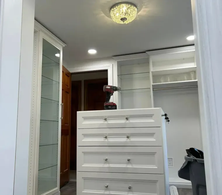 Walk-in closet with mirrored doors, white drawers, shelves, and a ceiling light in a gray-carpeted room