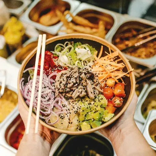 Handen die een kleurrijke poké bowl vasthouden met eetstokjes, gevuld met groenten, zeevruchten en sesamzaadjes.