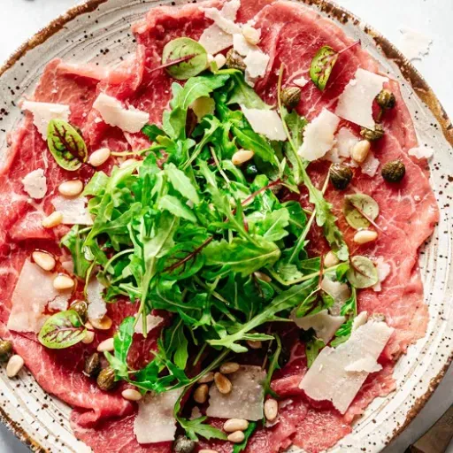 Rosé rundercarpaccio met rucola, geraspte Parmezaanse kaas en pijnboompitten op een wit bord.