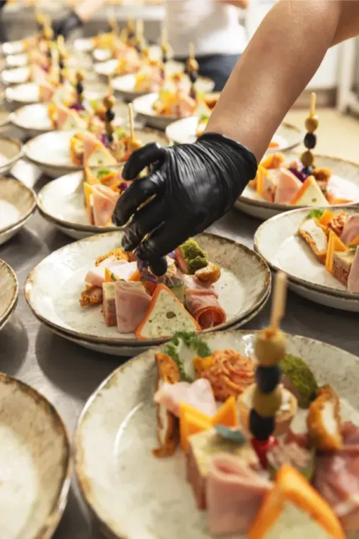 Een gehandschoende hand schikt kleine schaaltjes met hapjes, spiesjes en plakjes vlees op een feesttafel.