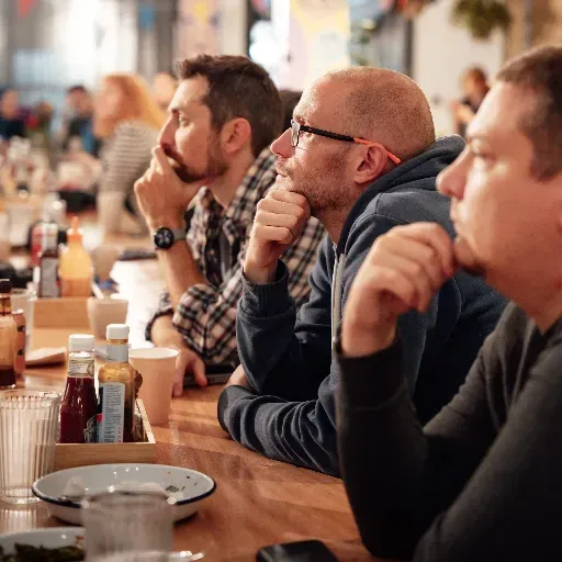 Mensen zitten aan een cafétafel en luisteren aandachtig, met hun drankjes en borden voor zich.