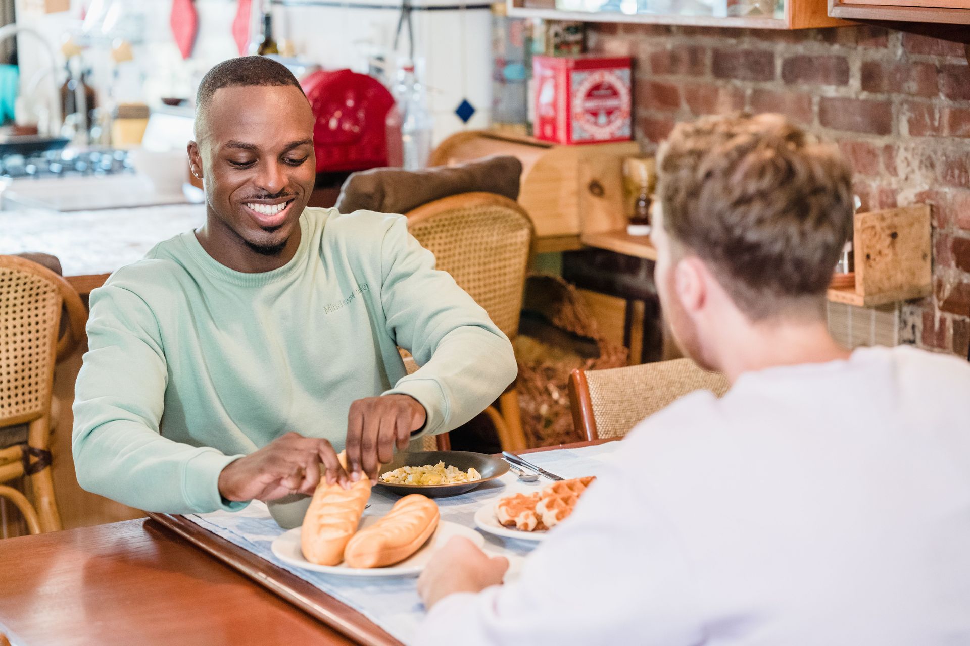 Twee mensen zitten aan een cafétafel te eten, glimlachend boven hun borden met eten en brood.